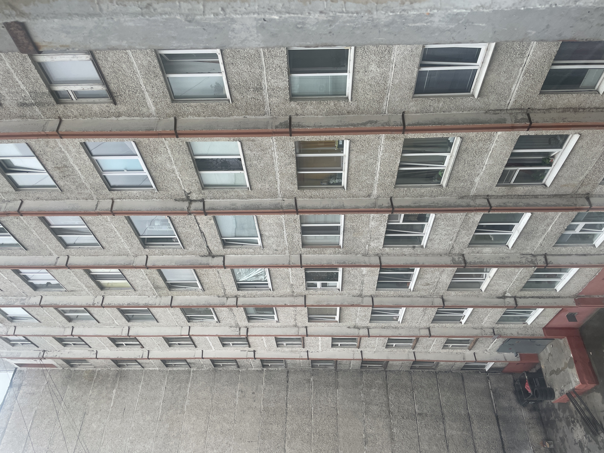 The image shows a tall, grey, concrete apartment building in Surgut, Russia. The building has numerous windows, many of which are white-framed and appear to be double-glazed. Some windows are open, while others are closed. Vertical red pipes run along the exterior of the building, attached by dark bands. On the ground level, there is an entrance with a grey door and a small concrete ramp with red trim. To the left of the entrance, there is a black dumpster with some red material inside and two tires stacked next to it. The overall appearance of the building and its surroundings suggests an older, utilitarian design common in residential areas of Russian cities. The sky is overcast, contributing to the dull, grey atmosphere. The image is taken from an elevated perspective, looking down at the building.