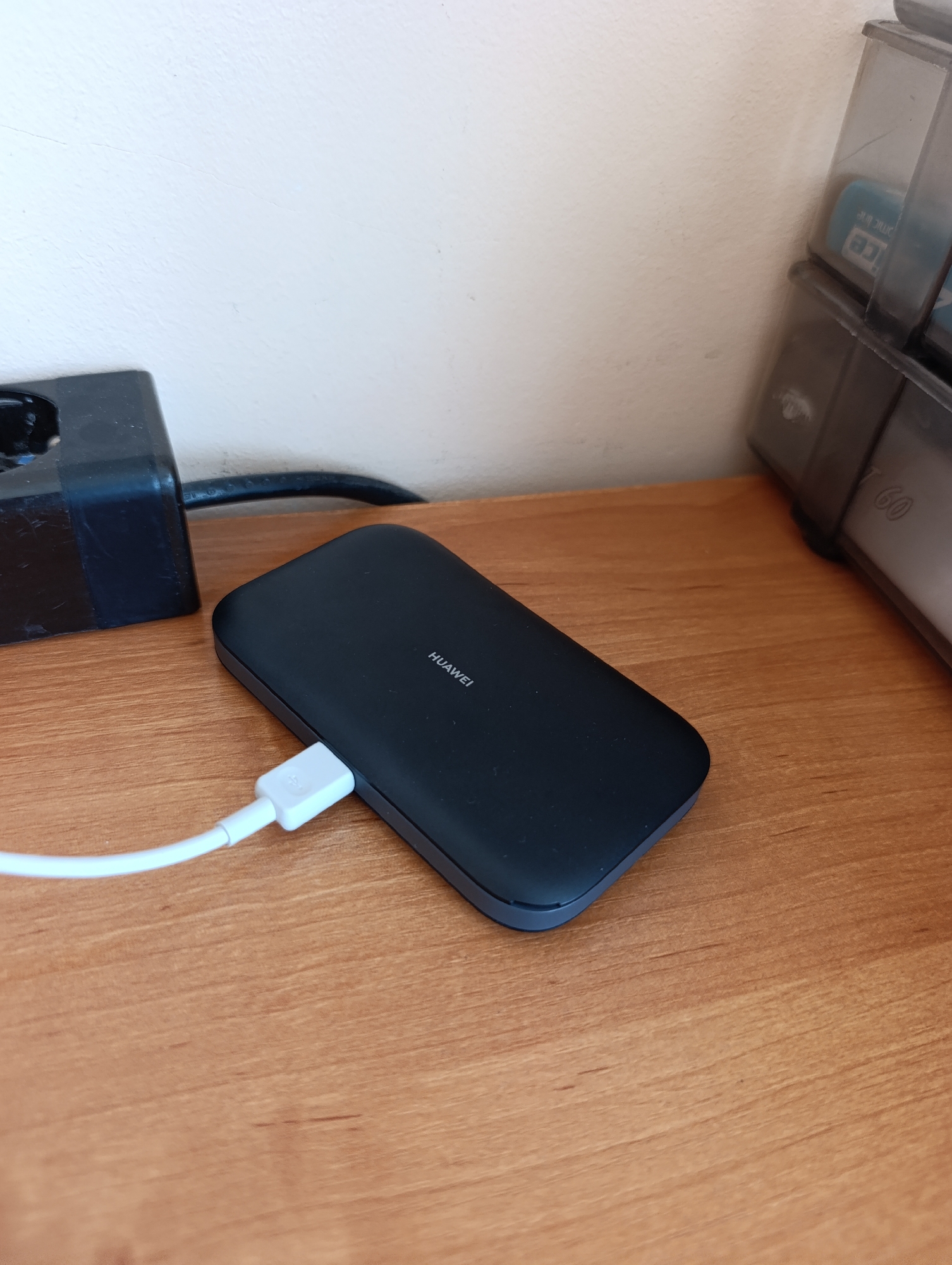 A black Huawei portable charger is connected to a white USB cable on a wooden desk. The cable is plugged into the charger, and the other end is out of frame. To the left of the charger is a black electrical adapter. In the background, a white wall and a clear plastic organizer containing blue items are visible. The scene is indoors.