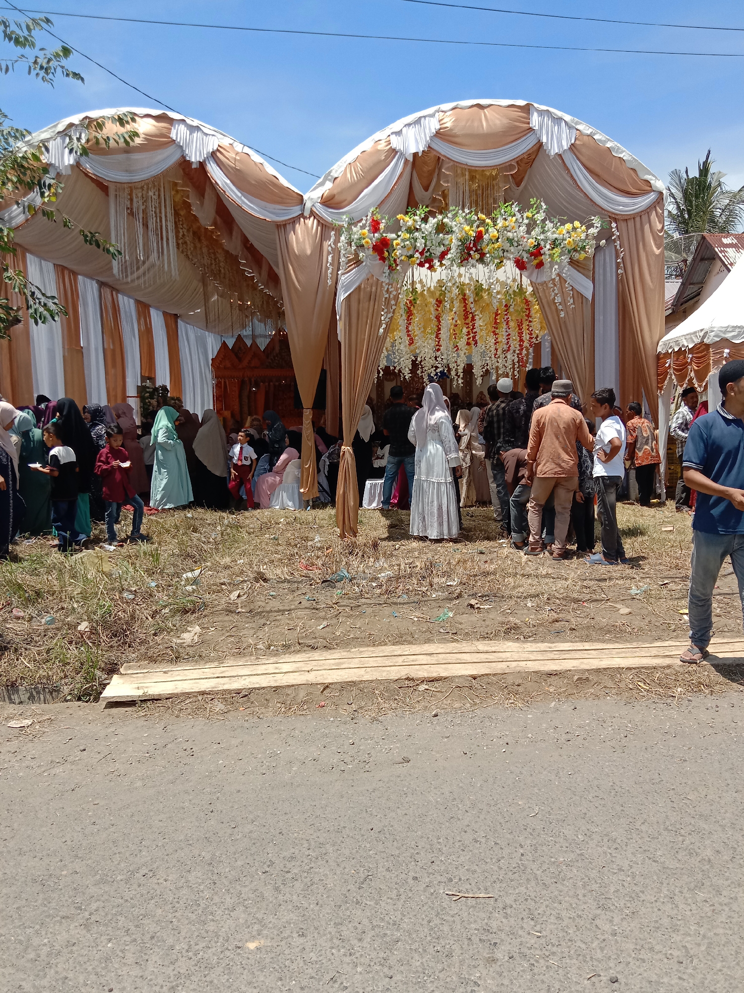 The image depicts an outdoor event, possibly a wedding or celebration, occurring in Lhokseumawe, Indonesia. A large tent or canopy structure is erected, decorated with fabric draping, floral arrangements, and hanging ornaments. A gathering of people is visible in and around the tent, consisting of men, women, and children. Some of the women are wearing hijabs. The people are interacting with each other, forming groups. The setting appears to be a roadside or open area with a mix of pavement, dirt, and some wooden planks. The weather is sunny, and the sky is clear.
