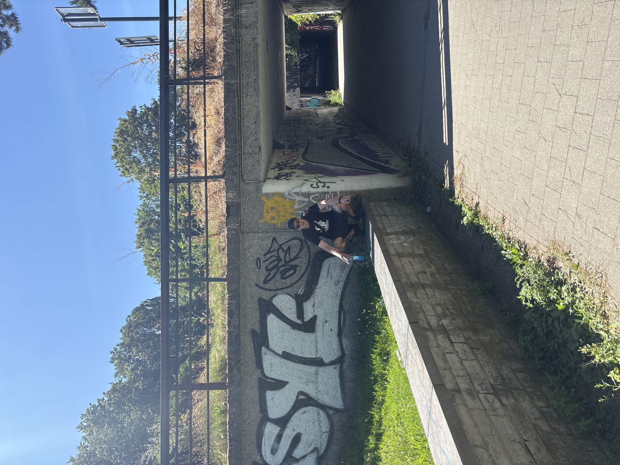 Two individuals are positioned beneath a concrete overpass adorned with graffiti. One person, wearing a black t-shirt, dark pants, sunglasses, and a black beanie, is seated on the ground. They are pointing towards something outside the frame with their right hand, while their left hand holds a light blue spray can. The other person is partially visible behind the seated individual, also seemingly engaged with the graffiti.

The scene is a passageway under a bridge, characterized by concrete structures and extensive graffiti artwork covering the walls. The ground is a mix of pavement and grassy areas. Sunlight casts shadows across the paved walkway, suggesting it is daytime. The sky is clear and blue, indicating fair weather.

Visible graffiti on the wall includes large, stylized lettering in white with black outlines, as well as other tags and drawings in various colors. The overall atmosphere appears relaxed and creative, with the individuals actively participating in or observing the graffiti art.

There is no visible text in the provided image. The location context provided is Brussels, Belgium, though this is not directly discernible from the visual elements of the image itself.