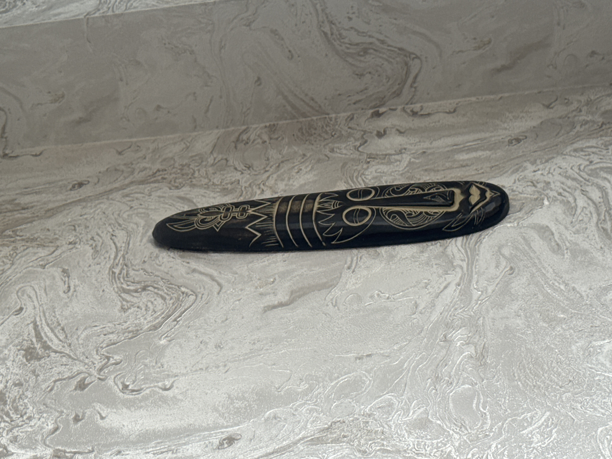 A dark, elongated, oval-shaped object with light-colored tribal or Polynesian-style carvings rests on a light gray and white marbled surface, possibly stone or tile. The background is a continuation of the same marbled surface, forming a wall or vertical plane. The object's detailed carvings are visible. The location is unknown; there is no information to suggest this is Tbilisi, Georgia.