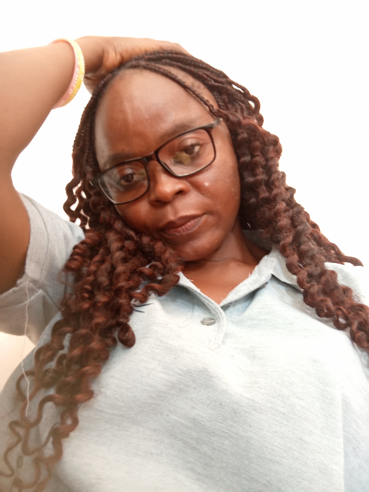 A close-up, slightly angled selfie features a Black woman wearing glasses and a light blue collared shirt. Her right arm is raised, with her hand resting on her head. A thin, multi-colored bracelet is visible on her wrist. Her hair is styled in dark brown braids, some of which fall over her shoulders and chest. The background is a plain, bright white, offering no specific location cues. The lighting suggests an indoor setting, possibly during the daytime. The woman's expression is neutral, and the overall mood of the image is casual and personal. There is no visible text in the image.