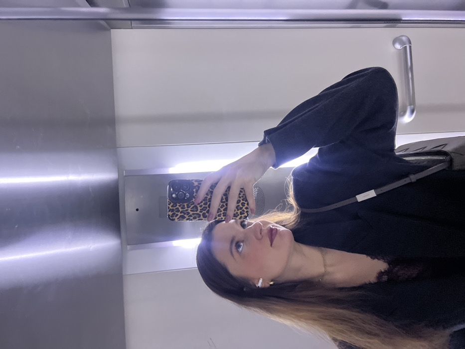 A woman with long brown hair, wearing a black blazer and a delicate lace top, takes a selfie in an elevator. She holds a leopard-print phone case. The elevator interior is sleek, with brushed metal walls and a handrail. Bright overhead lighting illuminates the scene, creating reflections on the metal surfaces. She wears a gold necklace and white wireless earbuds.