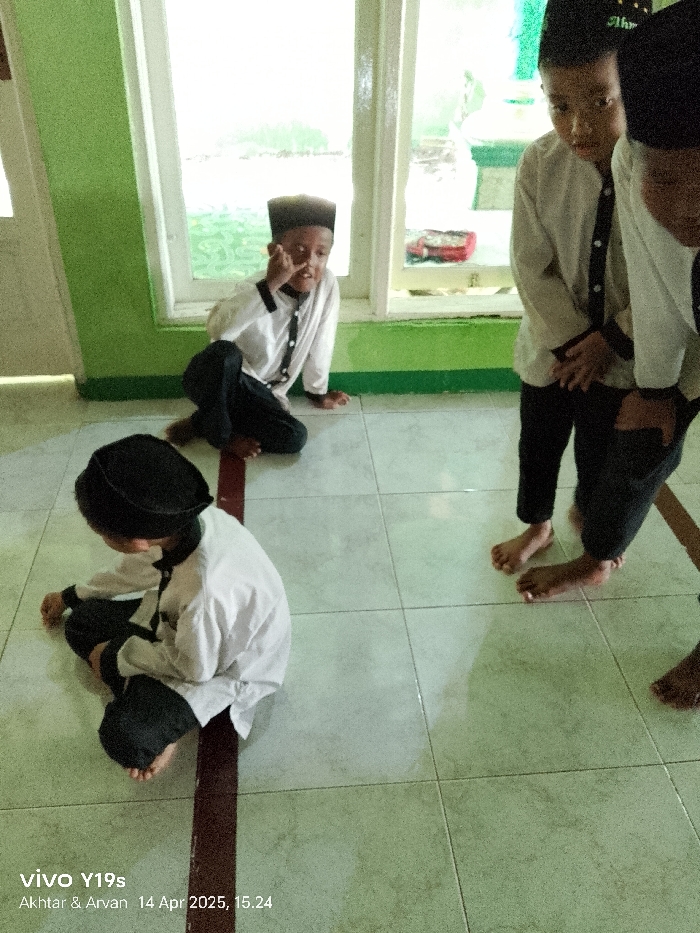 The image shows three young boys wearing traditional Islamic clothing in an indoor space with tiled flooring. The three boys are sitting on the floor, with one boy sitting on a burgundy line and another two standing further back. The boy on the line is wearing a black cap, a white shirt with a black buttoned jacket, and black trousers. The boy sitting behind him is wearing a black cap with a white top, and black trousers under a white shirt. The boy standing furthest away is wearing a black cap, a white shirt, and black trousers. The room is lit by natural light coming from a window on the right side of the image. The coordinates provided place this image in Anjatan, Indonesia, but without more context, it is impossible to pinpoint the exact location. The image appears to be taken inside a home or a school room.