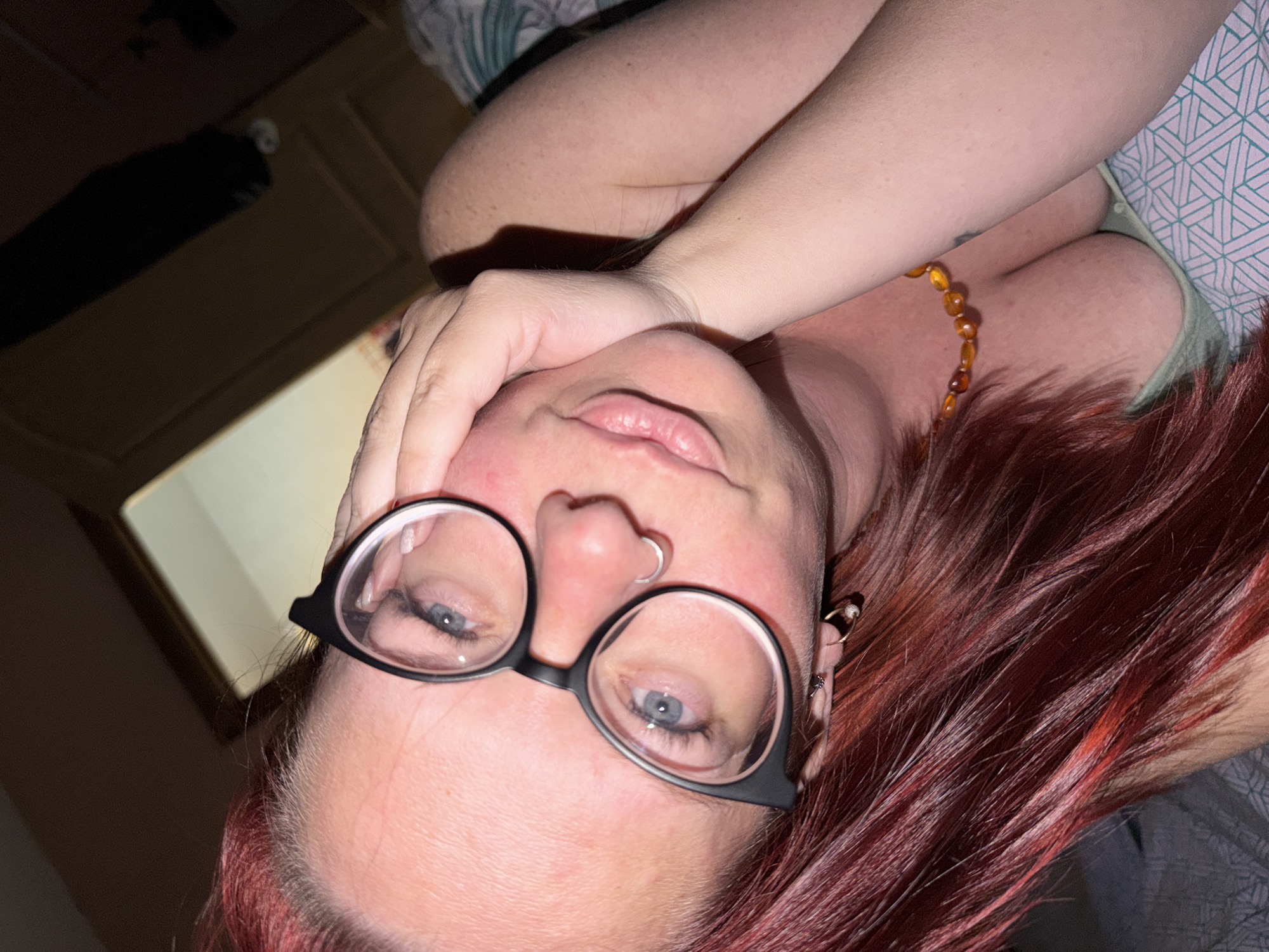 A selfie of a woman with reddish-brown hair wearing black-rimmed glasses is angled upwards towards the camera. She has fair skin and blue eyes. Her right hand is resting on her face. She has a nose ring and multiple earrings. She is wearing a necklace with amber-colored beads. The background includes a partially visible headboard and bedding. There is no indication of the location being Chaikovsky, Russia.