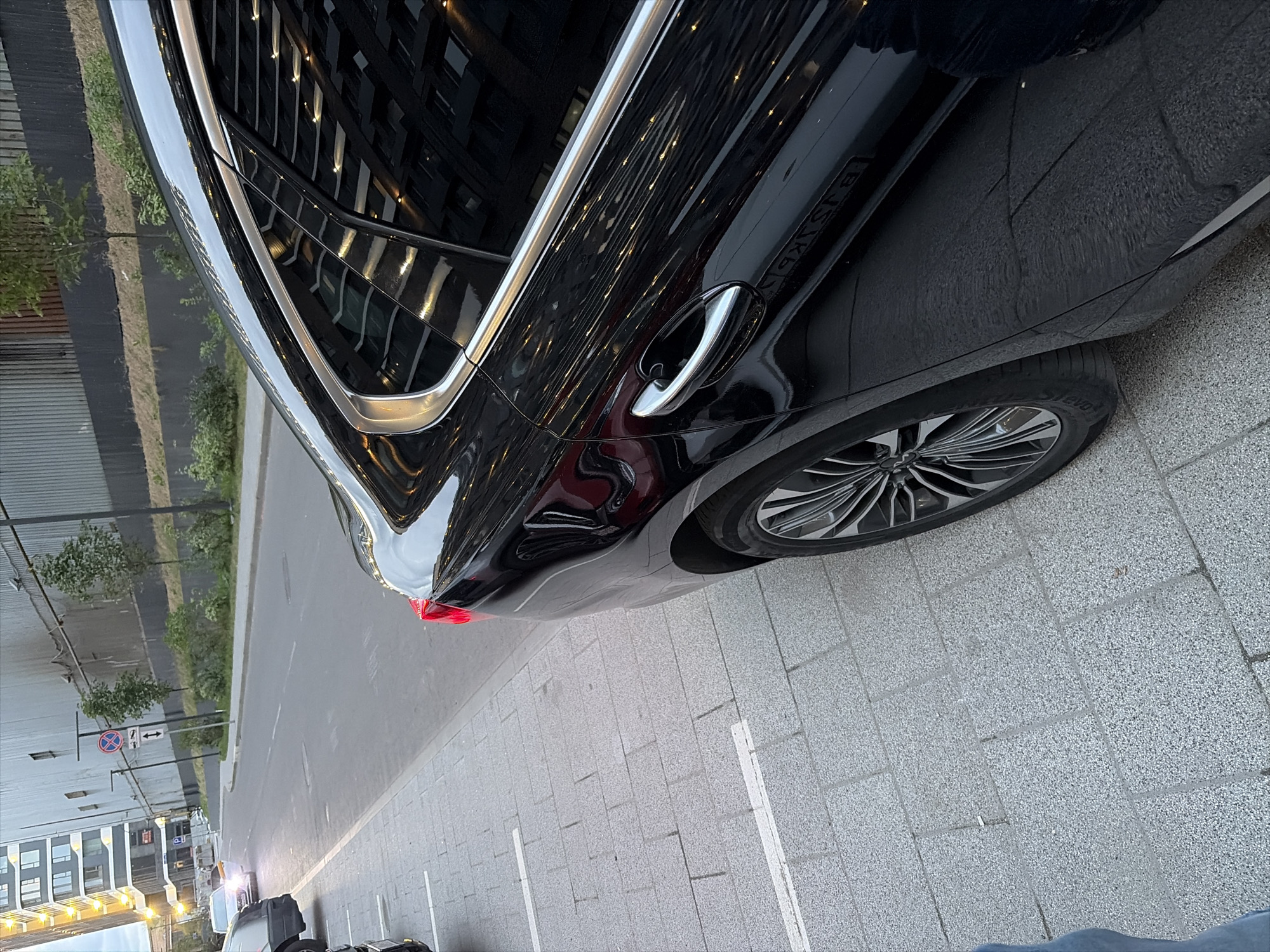 A black car is parked on a cobblestone sidewalk next to a street in a city. The car is facing away from the viewer, and only the rear half of the car is visible. The car has a chrome trim around the windows and a chrome door handle. The street is paved with asphalt and has a white line painted on it. There is a sidewalk on the other side of the street, and a building in the background. The building is tall and modern, with many windows. There are trees and bushes planted along the side of the street. The sky is overcast. The image was taken in Moscow, Russia.