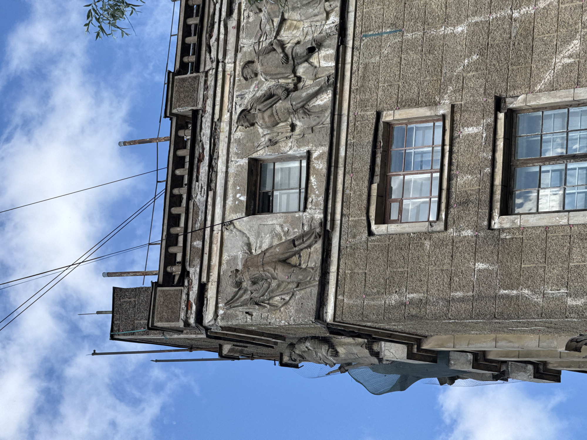 This image shows the facade of a building in Saint Petersburg, Russia, possibly undergoing renovation. The building features bas-relief sculptures of figures, some of which appear to be soldiers holding flags and rifles. The architecture is reminiscent of Soviet-era styles, characterized by its monumental and propagandistic art. The building's exterior shows signs of wear and tear, with crumbling plaster and weathered stone blocks. Several windows are visible, some with reflective glass that captures the sky and clouds. Overhead, electrical wires crisscross against a backdrop of a partly cloudy blue sky, and a branch of a tree is visible in the upper right corner. The building's corner is covered in green netting, suggesting ongoing construction or repair work.