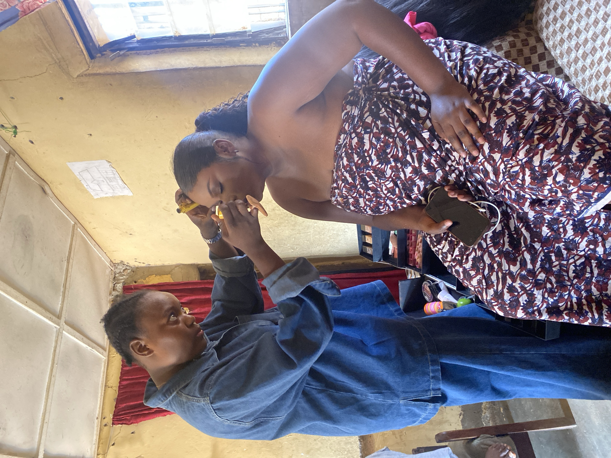In Panyam, Nigeria, a person in a denim jacket is applying makeup to another person who is seated and wrapped in a patterned fabric. The seated individual has their eyes closed and their face tilted upwards, while the person applying makeup holds a cosmetic tool near their eye. The environment appears to be an indoor space with yellow walls, a white ceiling, and red curtains visible in the background. There is also a window with a view of the outdoors. A makeup kit is open on a table beside the seated person, and a mobile phone rests on their lap.