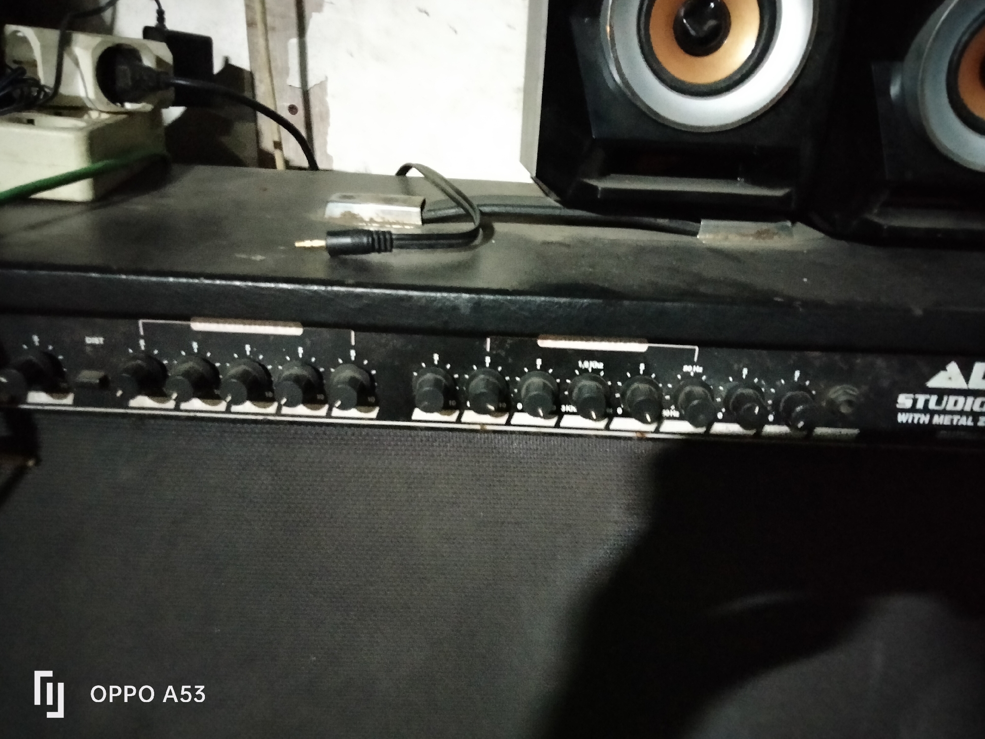 The image shows a close-up view of a black audio amplifier with multiple knobs and buttons on its front panel. There are two speakers visible at the top of the frame. A black audio cable with a 3.5mm connector is plugged into a silver adapter, which is resting on the amplifier's surface. In the background, a white electrical outlet with a black plug is visible, along with some cables. The overall environment appears to be indoors, with a slightly cluttered or unfinished appearance. The bottom left corner of the image shows a watermark "OPPO A53".