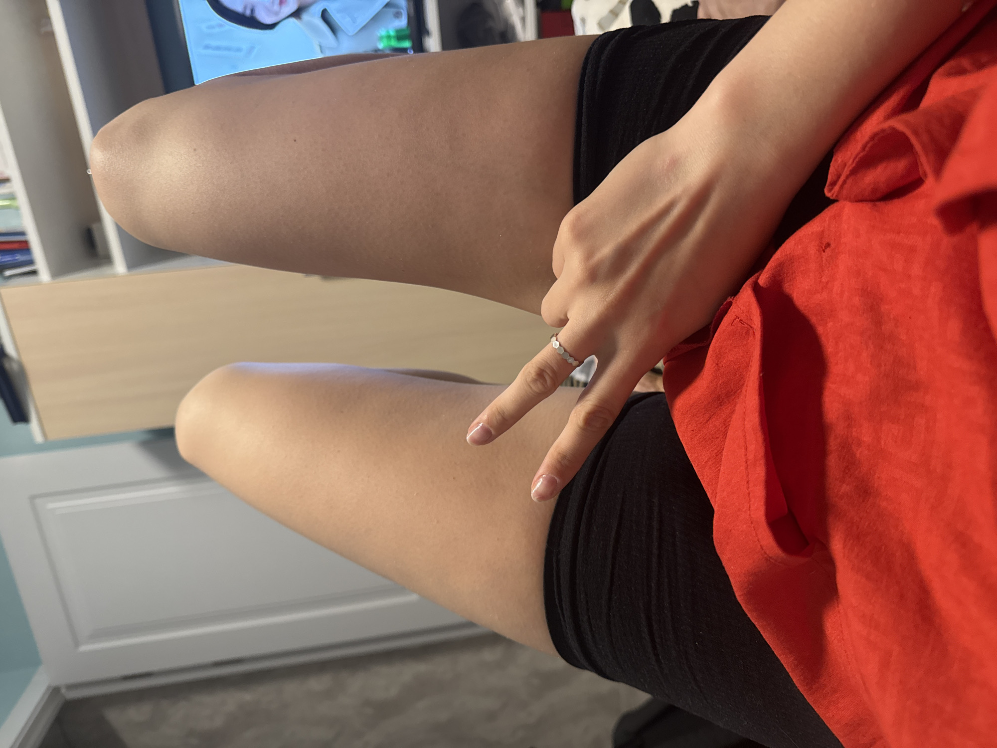 The image shows a person sitting with their legs crossed. They are wearing black shorts and a red shirt. Their hand is making a peace sign. A TV and furniture are visible in the background. The location is not identifiable as Izhevsk, Russia.