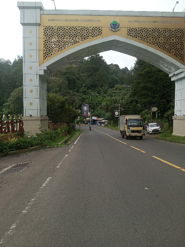 A gray asphalt road winds through a forest. An ornate archway, white and gold, frames the scene. A small yellow truck drives forward on the road. A white car is parked on the right. A motorcycle is visible in the distance. Lush trees cover the surrounding hills. The sky is overcast. The scene is bright overall, but not sunny.