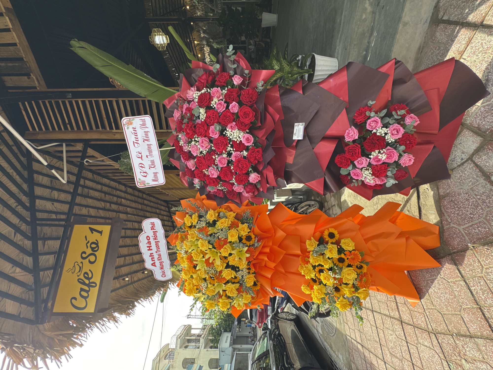 In Nha Trang, Vietnam, a street scene is captured with a prominent yellow sign reading "Cafe Số 1" hanging above a thatched roof structure. In front of the cafe, two large floral arrangements stand on display. The arrangement on the left is primarily composed of yellow roses and sunflowers, wrapped in bright orange paper. A smaller bunch of yellow roses is visible at the base. The arrangement on the right features a mix of red and pink roses, presented in a dramatic arrangement of red and dark brown wrapping. A small white tag is attached to this arrangement. In the background, a black sedan is parked on the left side of the frame. Behind the cafe, trees and more foliage are visible, along with hints of buildings and a glimpse of the sky. The ground is paved with patterned bricks.