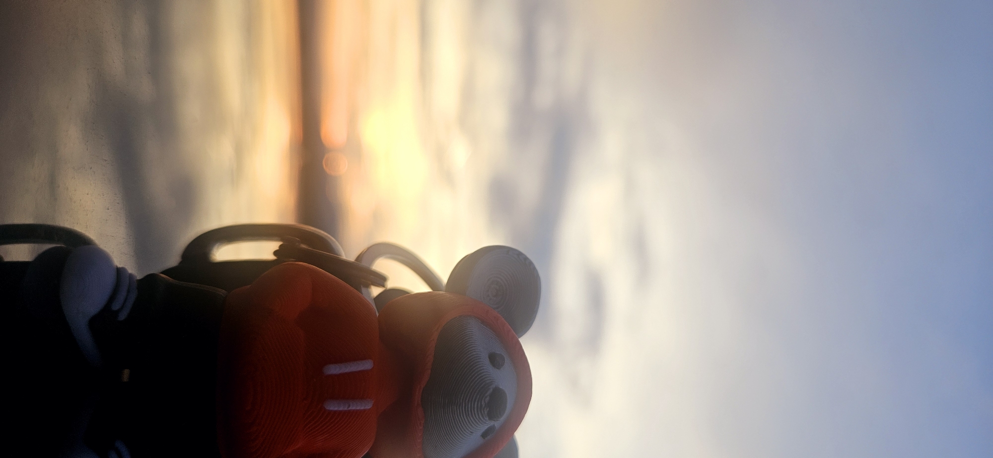 A close-up, vertical, low-angle shot focuses on a small, cartoonish figure wearing a red hoodie with white stripes, positioned in the foreground against a blurry, sunset-lit sky and body of water. The figure, which appears to be a mouse or similar character, is black with large, circular grey ears and a grey face with black eyes. It is attached to a grey keychain loop. The background features a gradient sky transitioning from pale blue at the top to warm orange and yellow hues near the horizon, with soft, diffused clouds. A calm body of water with reflections of the sky occupies the lower portion of the image. The overall scene has a soft focus and warm, ethereal lighting, suggesting an outdoor setting at dusk. There is no identifiable indication of the location being the city of Momin Prohod, Bulgaria.