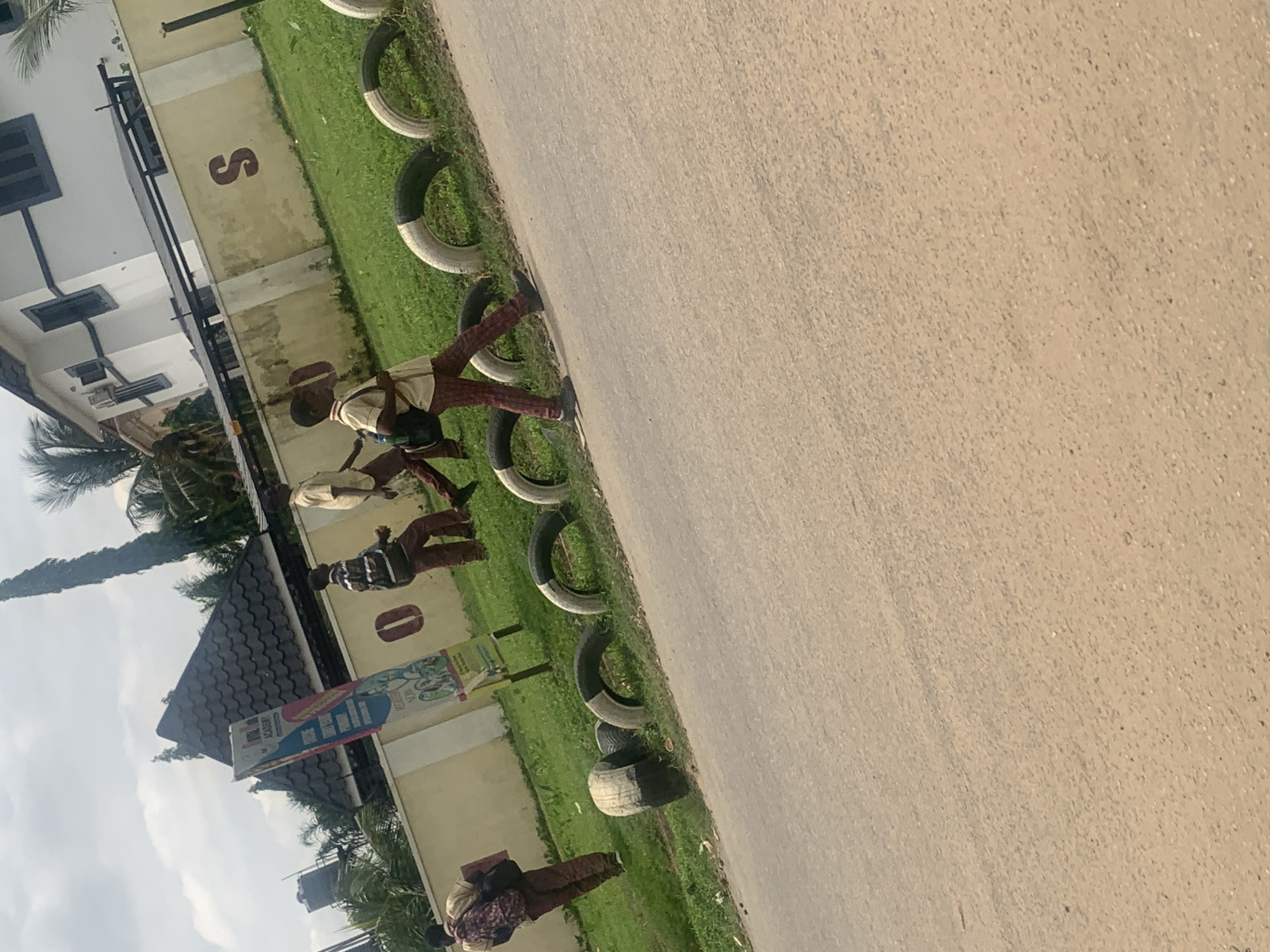 The image depicts an outdoor scene in Abuja, Nigeria, featuring a group of young people walking near a roadside. The environment includes a paved road in the foreground, a grassy area with several repurposed tires used as roadside barriers, and a wall behind that. A building, possibly a school or residence, with white walls and dark window frames, is visible in the background. A tall, slender tree stands to the left of the building. A large banner is posted on a pole near the wall.
There are at least four individuals, appearing to be students due to their attire, which includes light-colored shirts and patterned trousers. They are dressed in what seem to be school uniforms. One individual, on the far left, is seen from the back, carrying a backpack. Three other individuals are walking towards the right side of the frame. One of them is carrying a backpack on their shoulders. The individuals appear to be in motion, walking along the grassy area beside the road. The sky above is overcast with clouds. The overall impression is that of a typical urban or semi-urban street scene in Nigeria.