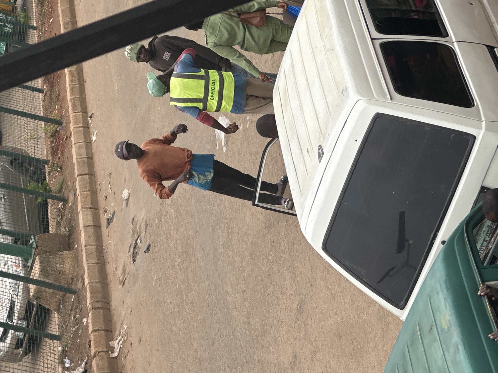 In Bukuru, Nigeria, a group of individuals, appearing to be either workers or bystanders, are gathered on a dusty, outdoor street. In the foreground, the roof of a white van is visible, obscuring the lower portion of the scene. To the left, the roof of a green vehicle is also present. The central focus of the image is on a group of four people in the middle ground. One man, wearing an orange long-sleeved shirt and dark trousers, stands facing the viewer with his hands visible. To his right, another man wears a bright yellow safety vest with the word "OFFICIAL" printed in black letters across the back. He has a green cap on and is wearing light-colored trousers. Behind him, another individual in a camouflage-patterned hat is partially visible. A fourth person, dressed in a green uniform, stands to the right of the man in the yellow vest, holding a brown bag. A small child is partially visible through an open car door. The background consists of a road surface, a concrete curb, and a chain-link fence. Beyond the fence, a car and some vegetation are discernible. The overall environment appears to be a somewhat unkempt urban setting.