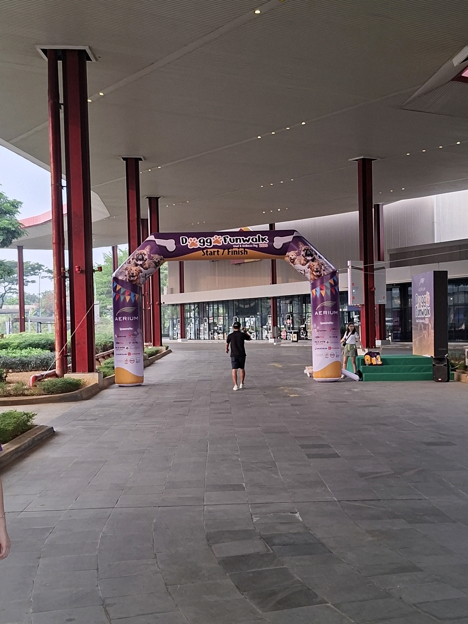 In Lengkong Kulon, Indonesia, an inflatable archway marks the "Dogg& Funwalk" start/finish line. The arch, decorated with images of dogs and sponsors' logos, stands on a paved walkway. A man wearing a black t-shirt and shorts walks away from the camera, heading towards the arch. To the right of the arch, a woman in a white t-shirt and olive-green shorts stands on a green tiered platform. A black speaker is placed on the ground next to the platform. In the background, the building features large windows and is supported by red pillars. Lush green trees and bushes are visible to the left of the walkway.