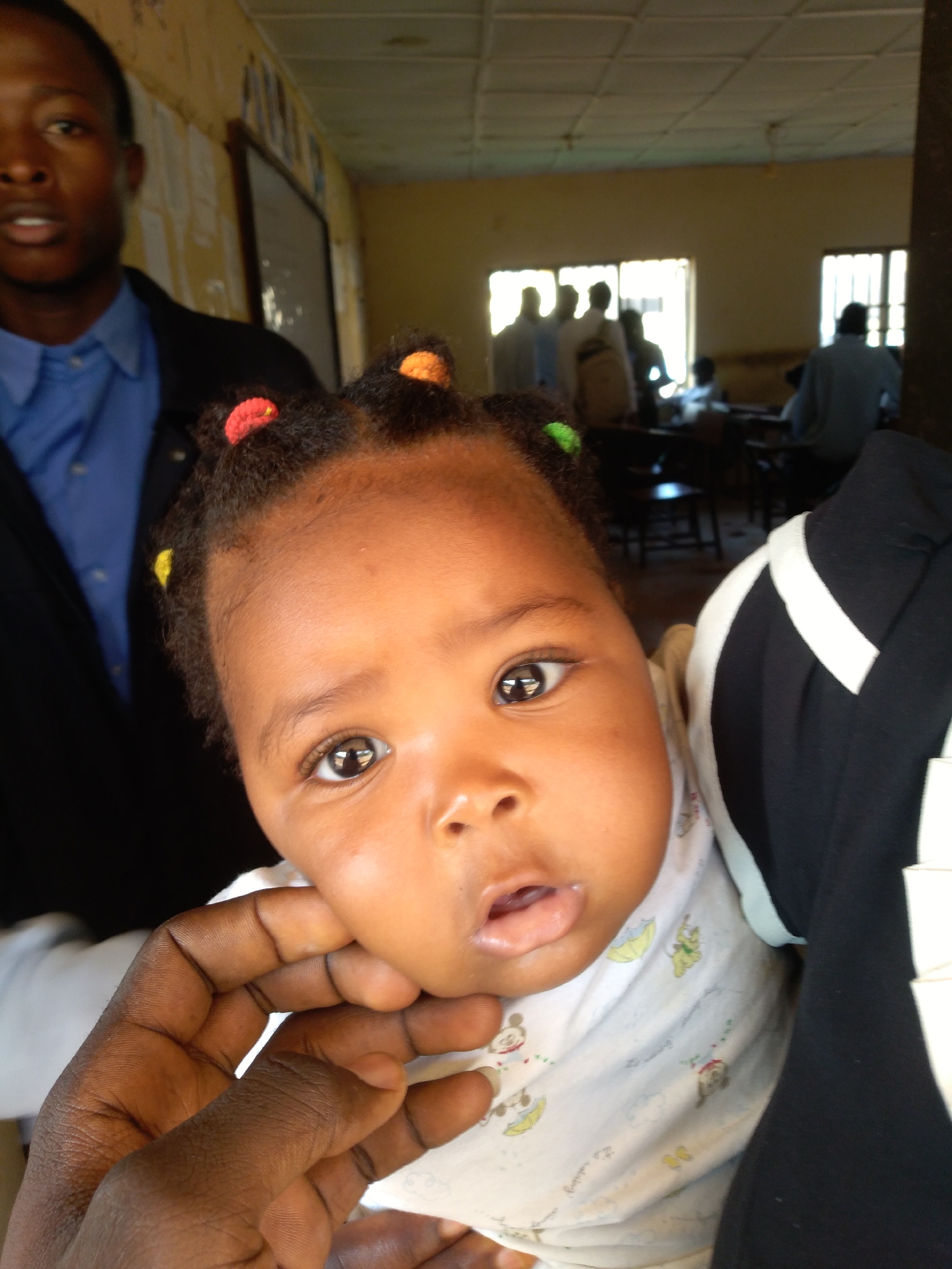 The image depicts a close-up of a dark-skinned baby's face, with the upper portion of an adult's face visible to the left. The baby has dark hair styled with colorful hair ties. The baby is looking directly at the camera, and its mouth is slightly open. An adult's hand is supporting the baby's chin. In the background, a room with tiled ceiling is visible, with several people silhouetted against windows. The overall impression is a candid, indoor shot, possibly in a community center or classroom.