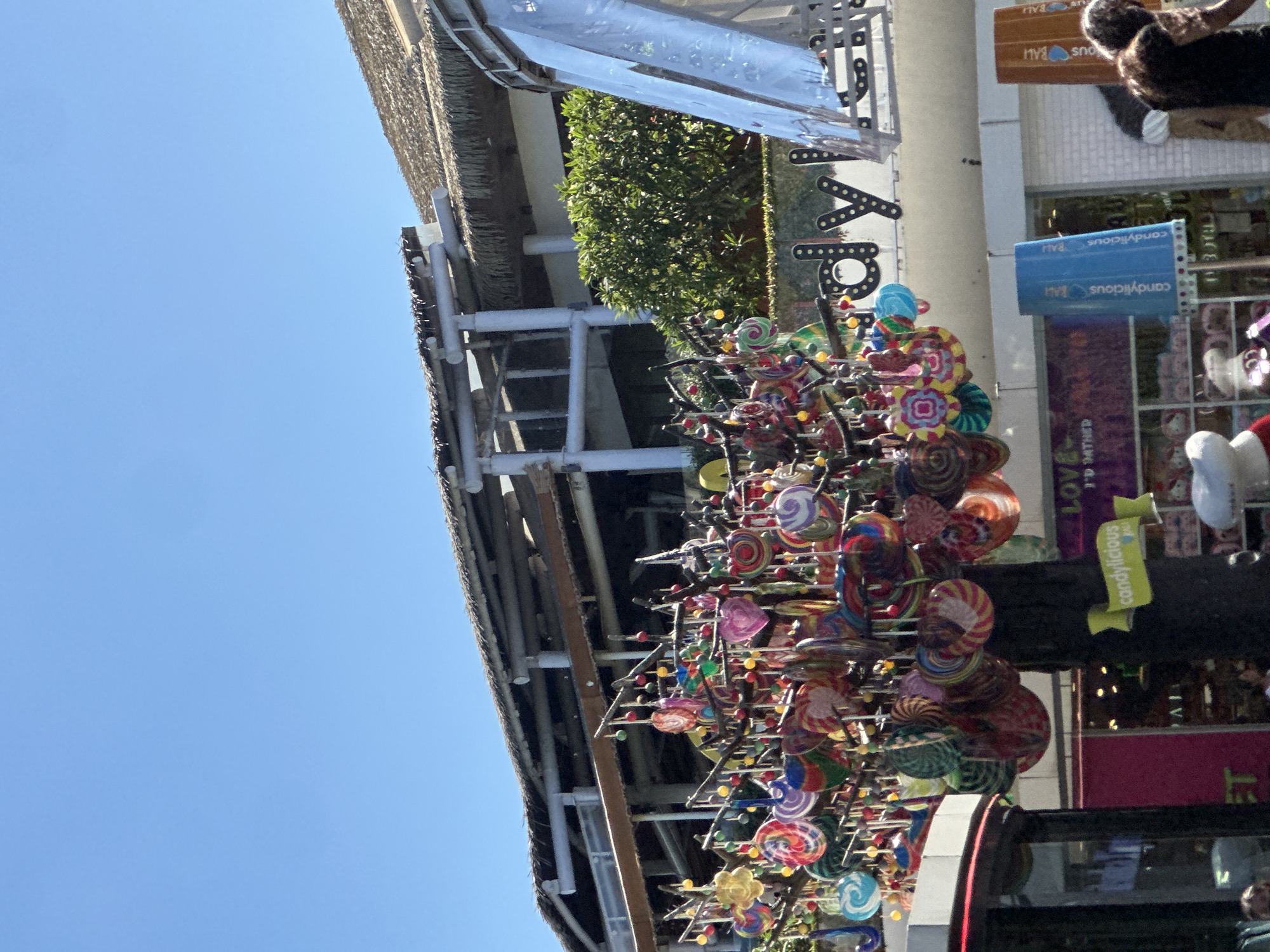 In Kuta, Indonesia, a tree adorned with colorful lollipops stands outside a store. The store's signage is visible, with large letters spelling out "candylicious". The building has a thatched roof and a modern architectural style. A blue cylindrical sign with "candylicious BALI" written on it is prominently displayed. In the background, there are shops and a clear blue sky. A person with long, curly hair is partially visible on the right side of the image.