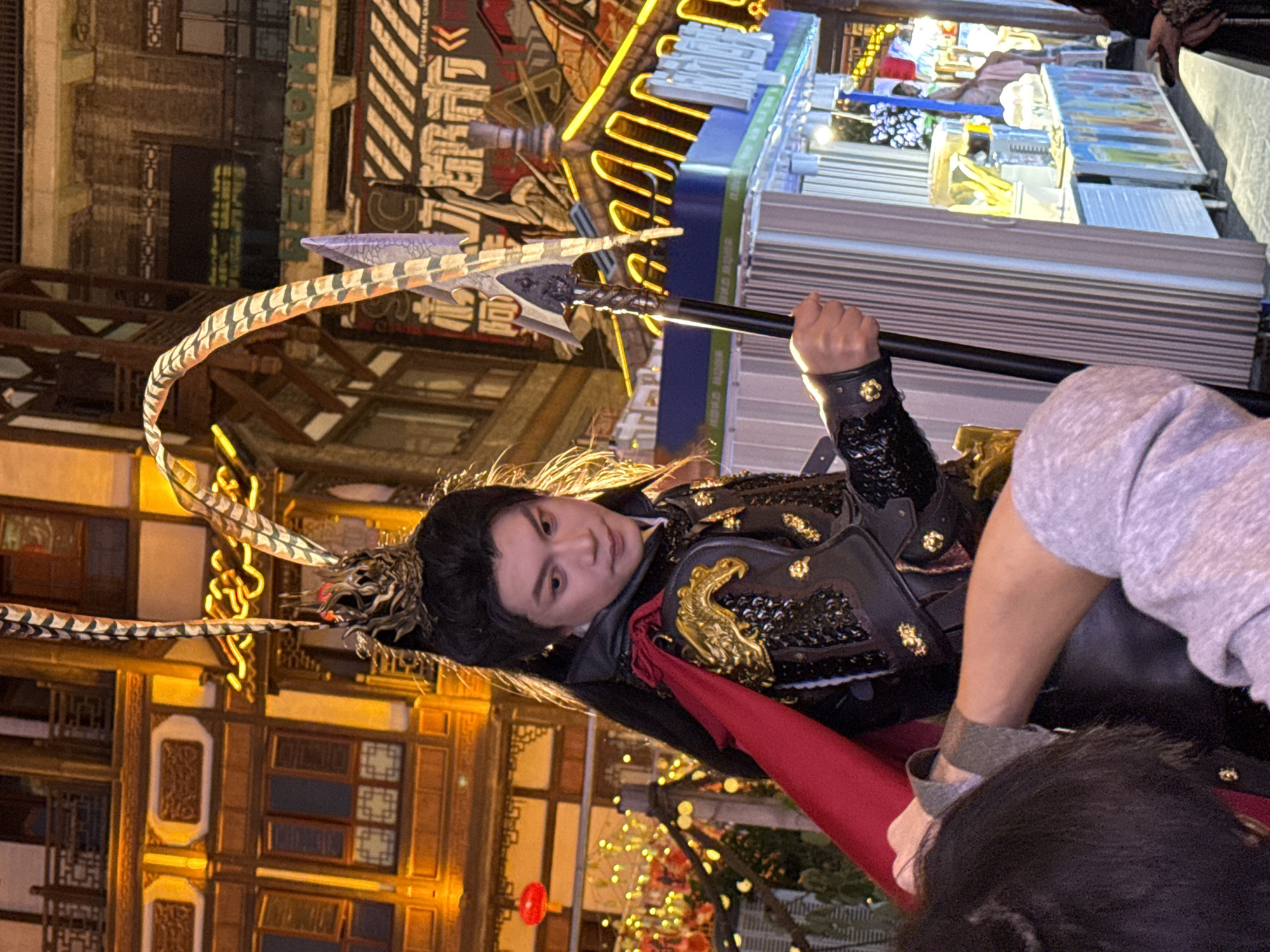 A person in ornate black and gold armor with a red cape stands holding a large, ornate spear. The person has dark hair pulled back and is looking towards the viewer. The background features a building with traditional Chinese architecture, as well as a street scene with various storefronts and neon lights. The location is Yuzhong District, China.