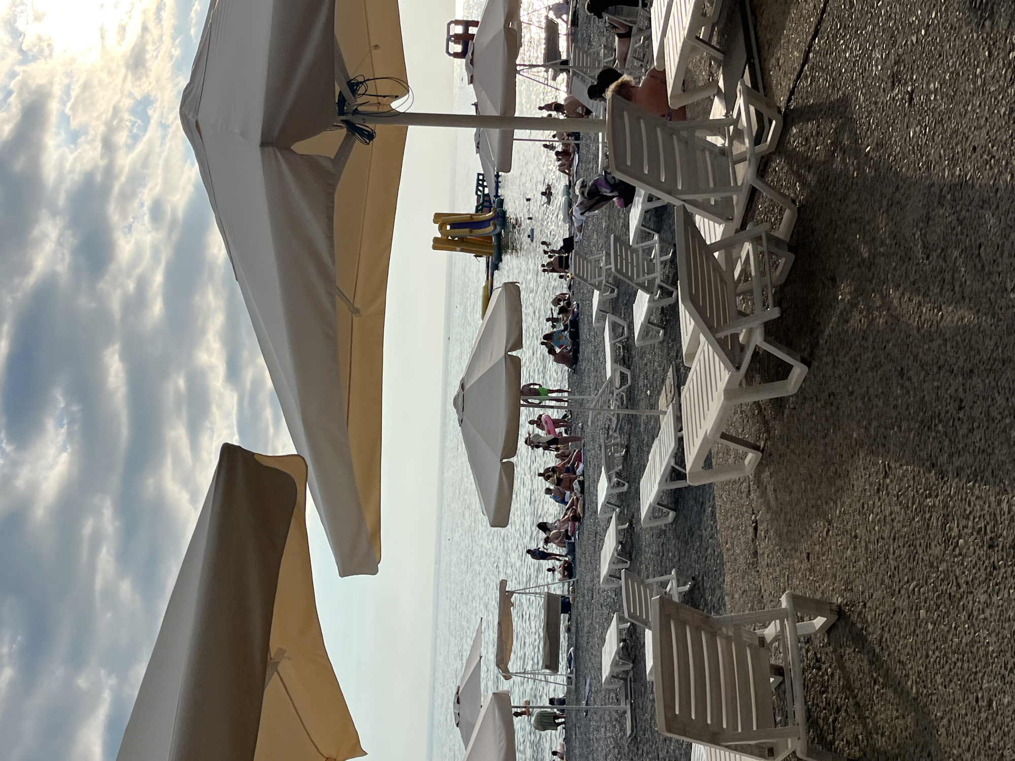 This is a photograph taken on a beach in Sochi, Russia. The scene depicts a lively beach with numerous people enjoying the water and sun.
The main subjects are the beachgoers, scattered across the pebble beach and in the shallow waters of the Black Sea. They are engaged in various recreational activities, including swimming, lounging on beach chairs, and playing. A yellow and blue inflatable water slide is visible in the sea, adding to the playful atmosphere.
Numerous white plastic lounge chairs are set up on the beach, many occupied by people or casting shadows. Several large beige beach umbrellas provide shade, with two prominently in the foreground, partially obscuring the view. The sea is calm, reflecting the bright sky.
The time of day appears to be late afternoon or early evening, as indicated by the quality of light and the long shadows cast by the chairs and umbrellas. The weather seems pleasant, with a partly cloudy sky where sunlight breaks through, creating a somewhat dramatic effect. The pebbly texture of the beach is a characteristic detail of many Black Sea coastlines. The overall mood is one of relaxation and enjoyment.
No visible text is present in the image.