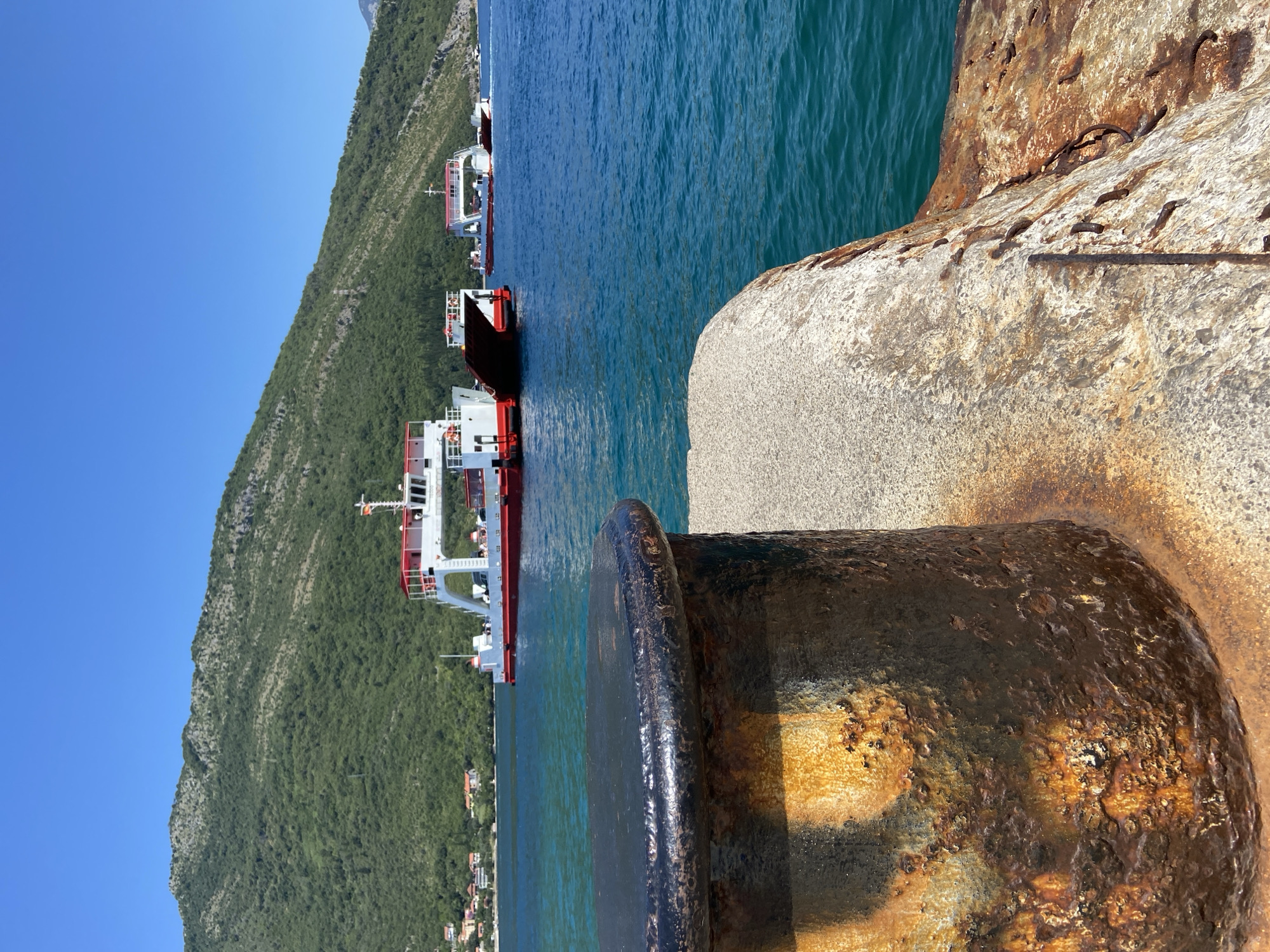 In Lepetane, Montenegro, a sunny day unfolds with ferries operating on a clear blue sea. In the foreground, a large, rusted bollard sits on a concrete dock. Behind it, the turquoise water stretches towards the horizon. Three white and red ferries are visible in the middle ground. The largest ferry, positioned closest to the viewer, has its ramp lowered. Another ferry is further out, and a third is in the distance. Lush green mountains, dotted with houses, form the backdrop, under a bright blue sky.