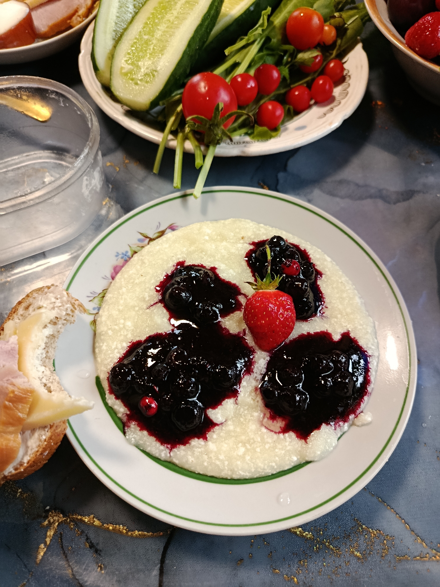 A close-up shot captures a plate of white porridge topped with blueberry sauce and a single strawberry. To the left, a partially eaten sandwich with ham and cheese rests on a dark, textured surface. In the background, a plate of sliced cucumbers and cherry tomatoes is visible, along with a bowl of strawberries. The scene is set on a table with a blue and gold marbled finish.