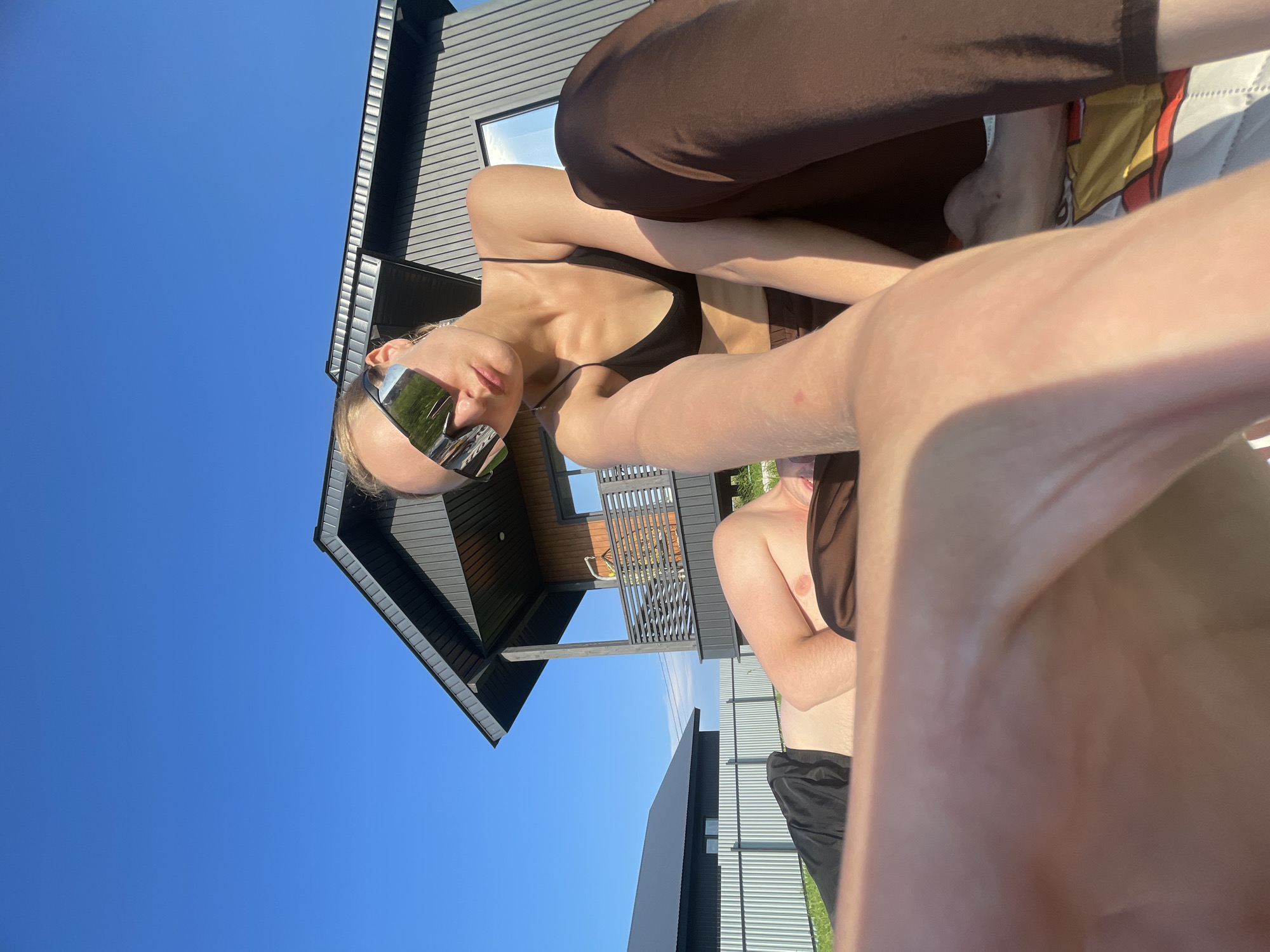 A young woman with blonde hair, wearing futuristic mirrored sunglasses and a black bikini top, is pictured outdoors. She is lying down, with her legs crossed and her arm extended towards the camera, creating a selfie-like perspective. Beside her, another person, a young man, is also visible, lying down and wearing black shorts. They are on a white blanket with a colorful pattern, possibly on a patio or lawn. In the background, a modern dark gray house with a steep roofline and wooden accents is seen. The clear blue sky suggests a sunny day. The overall impression is one of leisure and relaxation in a suburban or rural setting, possibly during the daytime in summer.