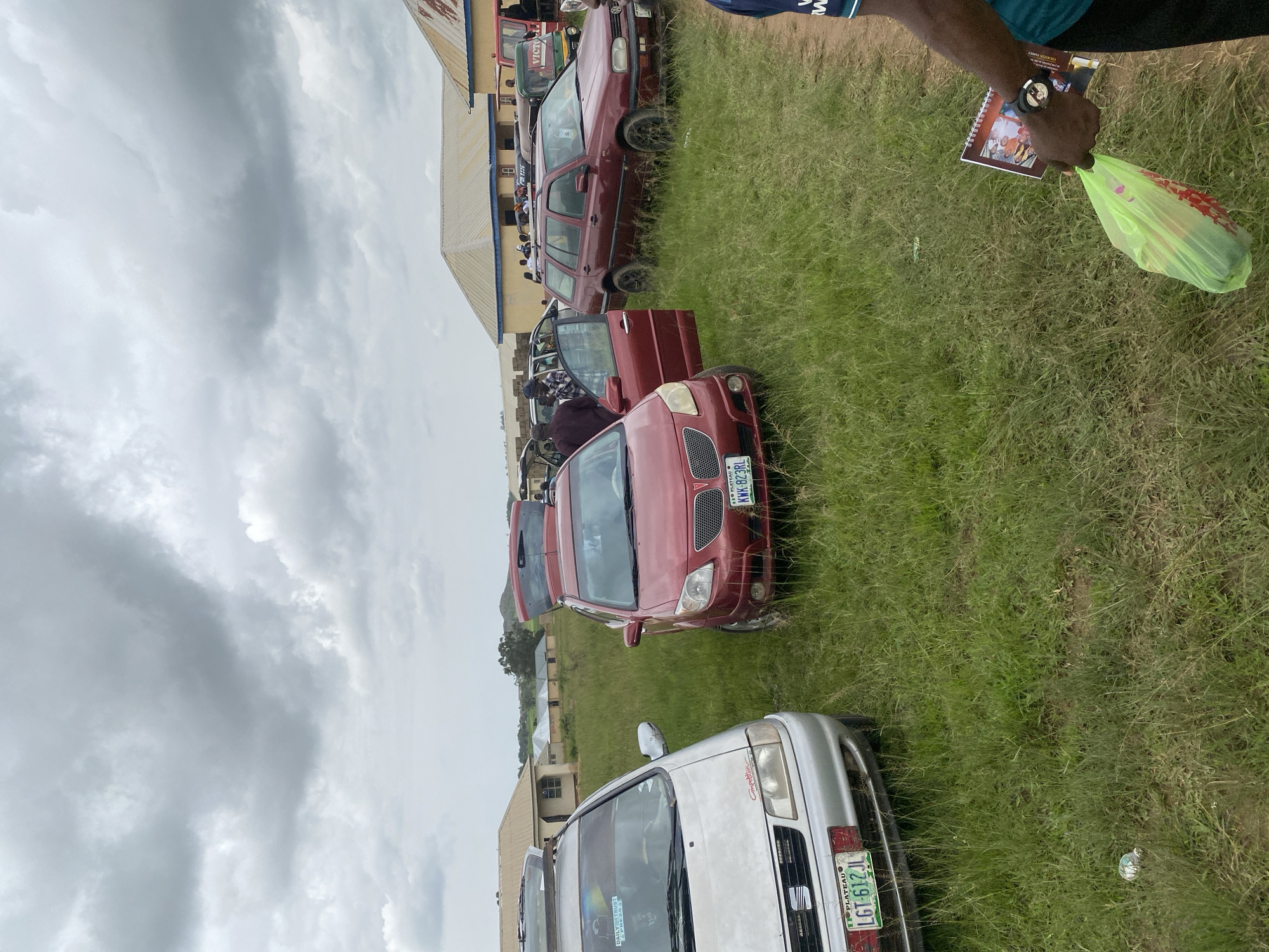 The image depicts an outdoor scene in Durumi, Nigeria, characterized by a cloudy sky and a grassy field with several vehicles. A silver sedan is parked on the left, with its hood open, and a red hatchback is positioned to its right, also with its trunk and driver's side door open. A maroon station wagon is further to the right, with its rear hatch open. Several people are visible near the vehicles, some appearing to be engaged in activities around the cars. In the background, there are buildings with light-colored roofs, suggesting a semi-urban or rural setting. A person in the foreground on the right is holding a green plastic bag and a booklet. The overall impression is one of a gathering or event where vehicles are being utilized for transport or storage.
