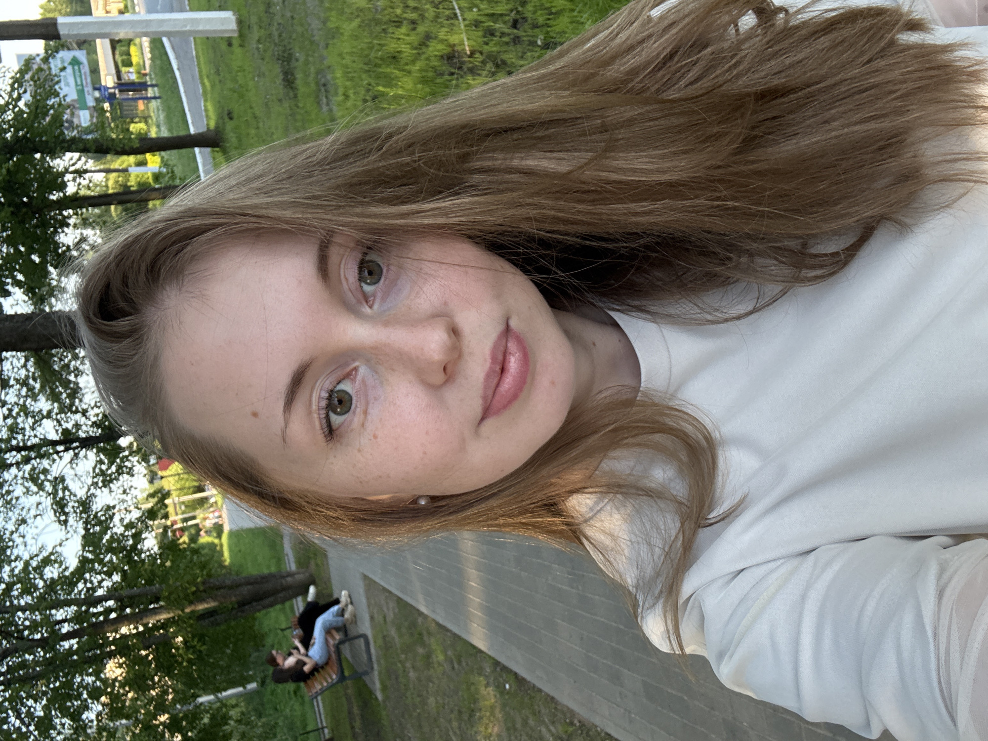 A young woman with light brown hair and fair skin takes a selfie in a park. She is wearing a white top. In the background, a paved walkway runs through a grassy area with trees. Two people are sitting on a bench in the distance.