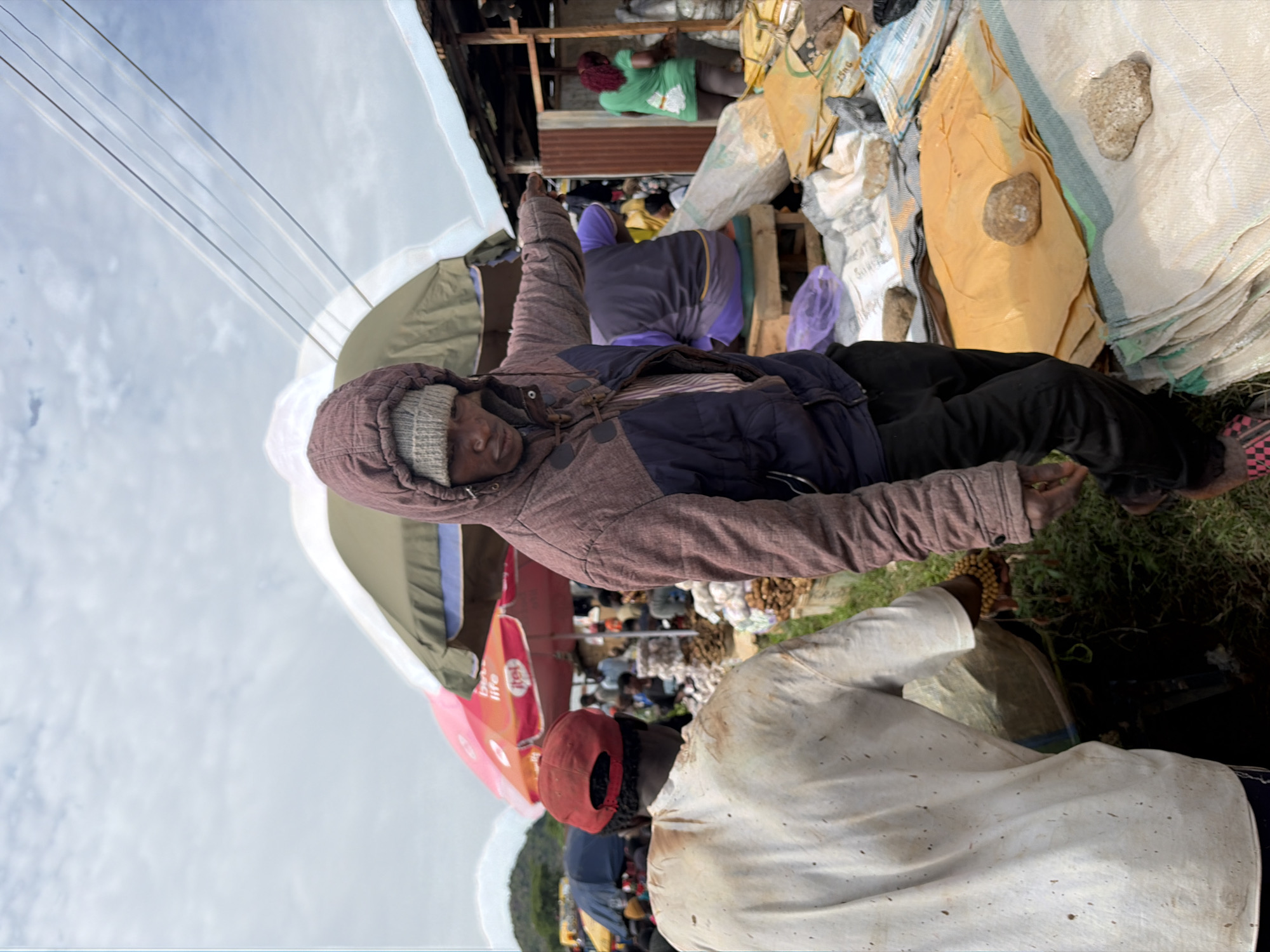 In Gura Top, Nigeria, an outdoor market scene unfolds under a cloudy sky. In the foreground, a person wearing a hooded jacket and a beanie gestures towards the left side of the frame. To their left, another individual in a dirty white shirt and a red cap is partially visible, facing away from the camera and holding a bag or sack. The market is filled with various stalls, indicated by colorful umbrellas and makeshift structures. Piles of goods, including sacks of produce and other items, are displayed on the ground, some covered with yellow and white tarps. Several other people are present in the background, engaged in market activities. Overhead, power lines are strung across the sky. The overall environment suggests a bustling and active marketplace.