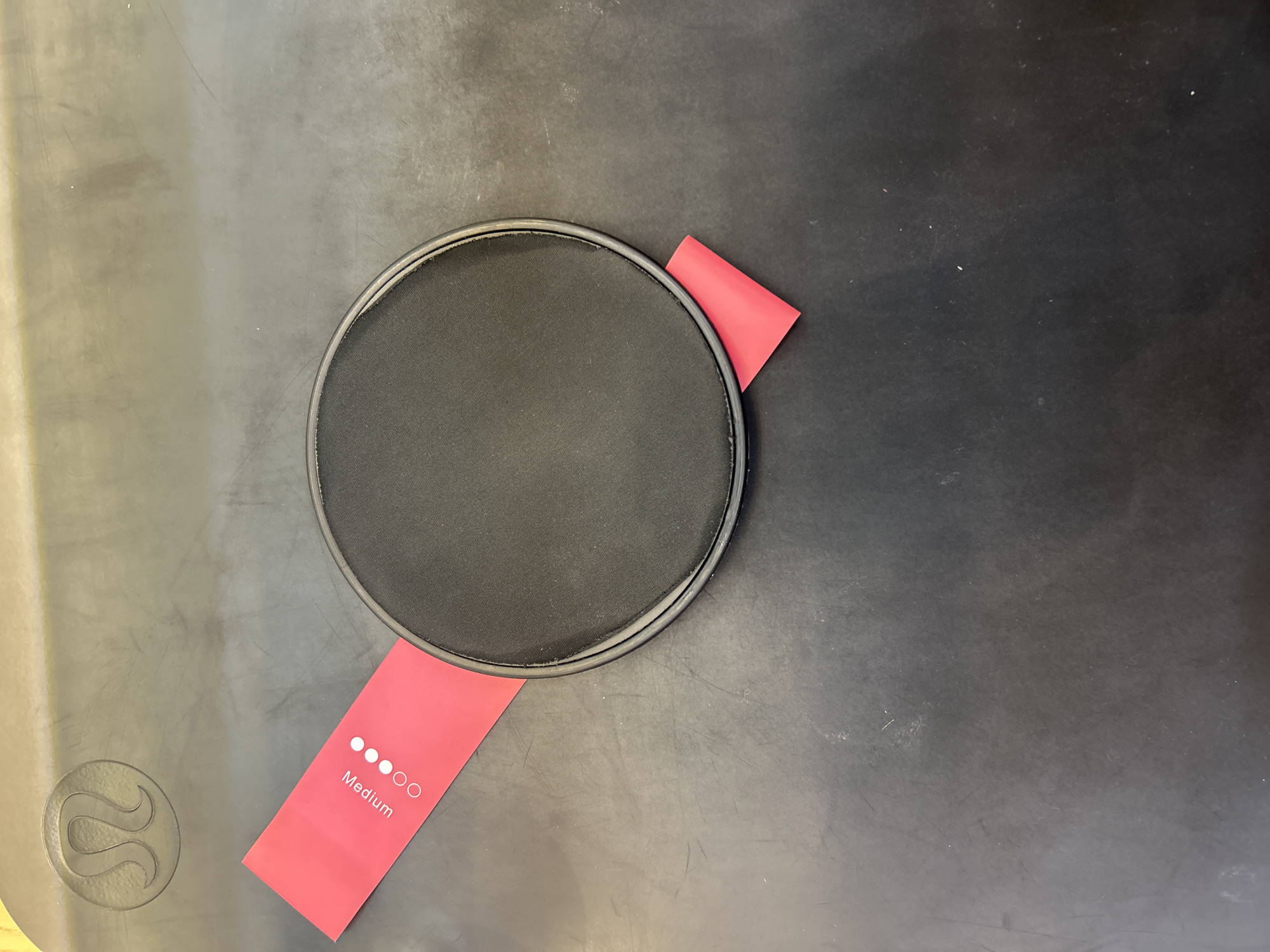 The image displays a flat lay composition of fitness equipment on a dark grey or black mat. The central subject is a circular, black fitness glider or sliding disc, featuring a slightly textured, fabric-like top surface and a dark rim. Partially underneath and extending from the glider is a bright red resistance band.
Notable details include text on the red resistance band, which clearly reads "Medium," accompanied by five dots above it. Three of these dots are filled in white, while the remaining two are empty outlines, likely indicating the resistance level. In the bottom left corner of the mat, a faint, embossed logo resembling the Lululemon symbol (a stylized 'A' or swoosh) is visible. The mat itself appears to be made of rubber or a similar material, showing some minor scuff marks and dust particles, suggesting use. No activity is taking place; the items are stationary. The setting appears to be an indoor space, possibly a home gym or fitness studio, given the nature of the equipment and mat.