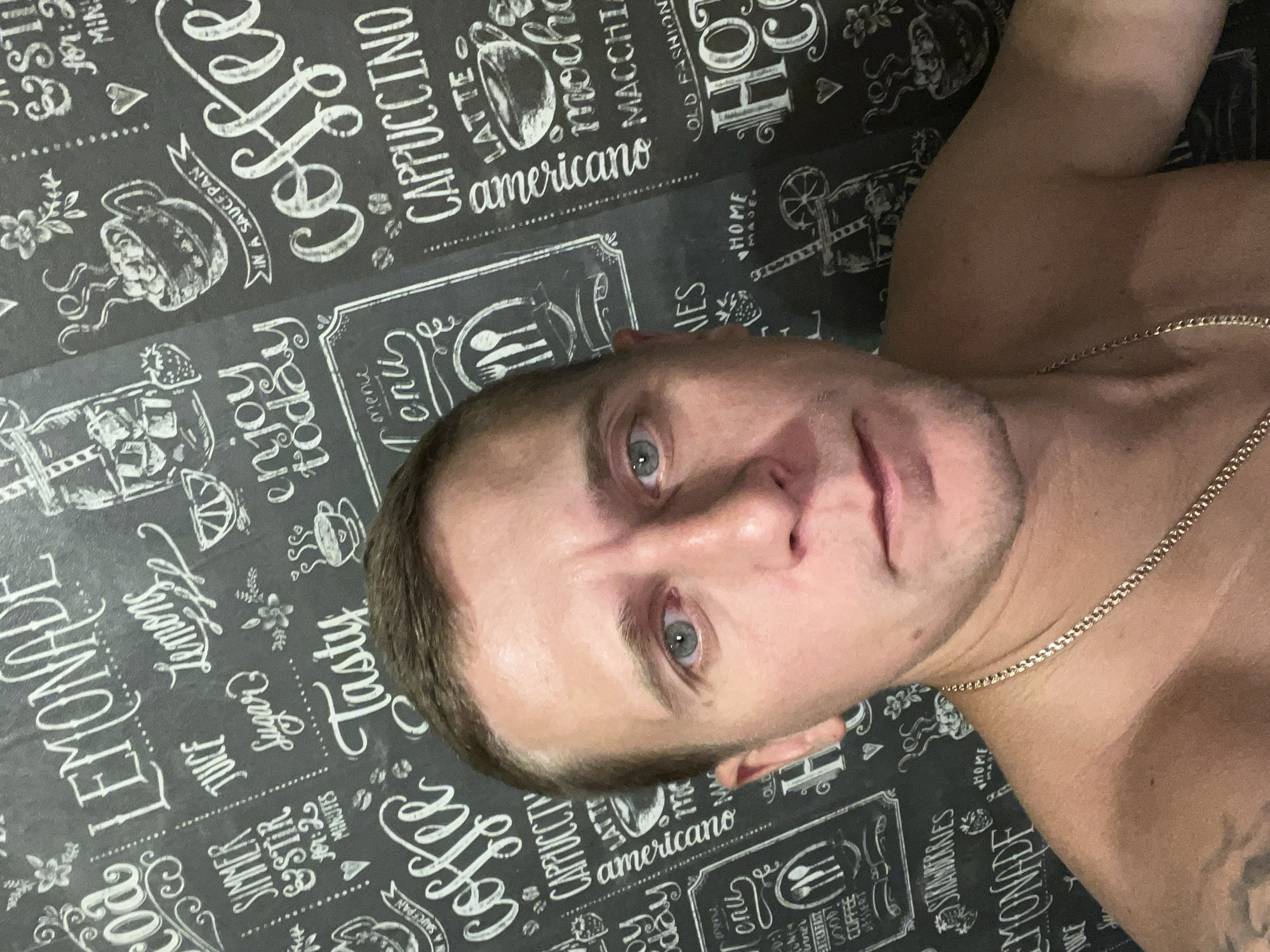 A close-up, low-angle, outdoor selfie of a white male in his late 30s or early 40s is shown against a black chalkboard-style wallpaper featuring various coffee and lemonade-themed drawings and text. The man is shirtless, wearing a gold chain around his neck. His hair is short and brown, and he has blue eyes. He has a fair complexion and appears to be clean-shaven. His left arm and shoulder are visible, with a tattoo on his left bicep. The wallpaper behind him is decorated with illustrations of coffee cups, fruit, and text in white cursive and block letters, including "LEMONADE," "COFFEE," "CAPPUCCINO," "LATTE," "AMERICANO," "JUICE," and "STRAWBERRIES." The background suggests an indoor setting, possibly a cafe or kitchen, with no discernible urban elements indicating a specific location in Minsk, Belarus. The lighting is flat, and the image appears to have been taken with a flash.
