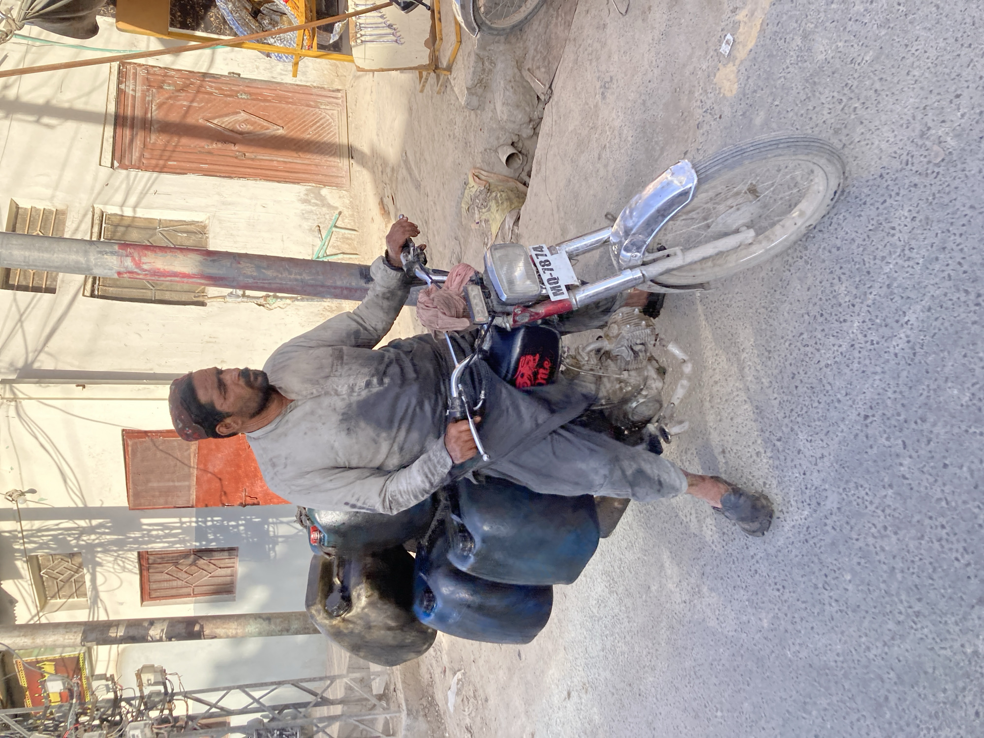 The image depicts a man seated on a motorcycle parked on a paved surface, likely a street. The man, of South Asian descent, wears a light-colored, long-sleeved shirt and darker pants. He is facing towards the left of the frame, holding the motorcycle handlebars. The motorcycle is laden with what appear to be several containers. The license plate of the motorcycle reads "MQ-7874".
The background features buildings with bricked window frames and doors, suggesting an urban setting. Electrical poles and wires are visible in the periphery. Based on these elements, the location is consistent with Quetta City Tehsil in Pakistan.