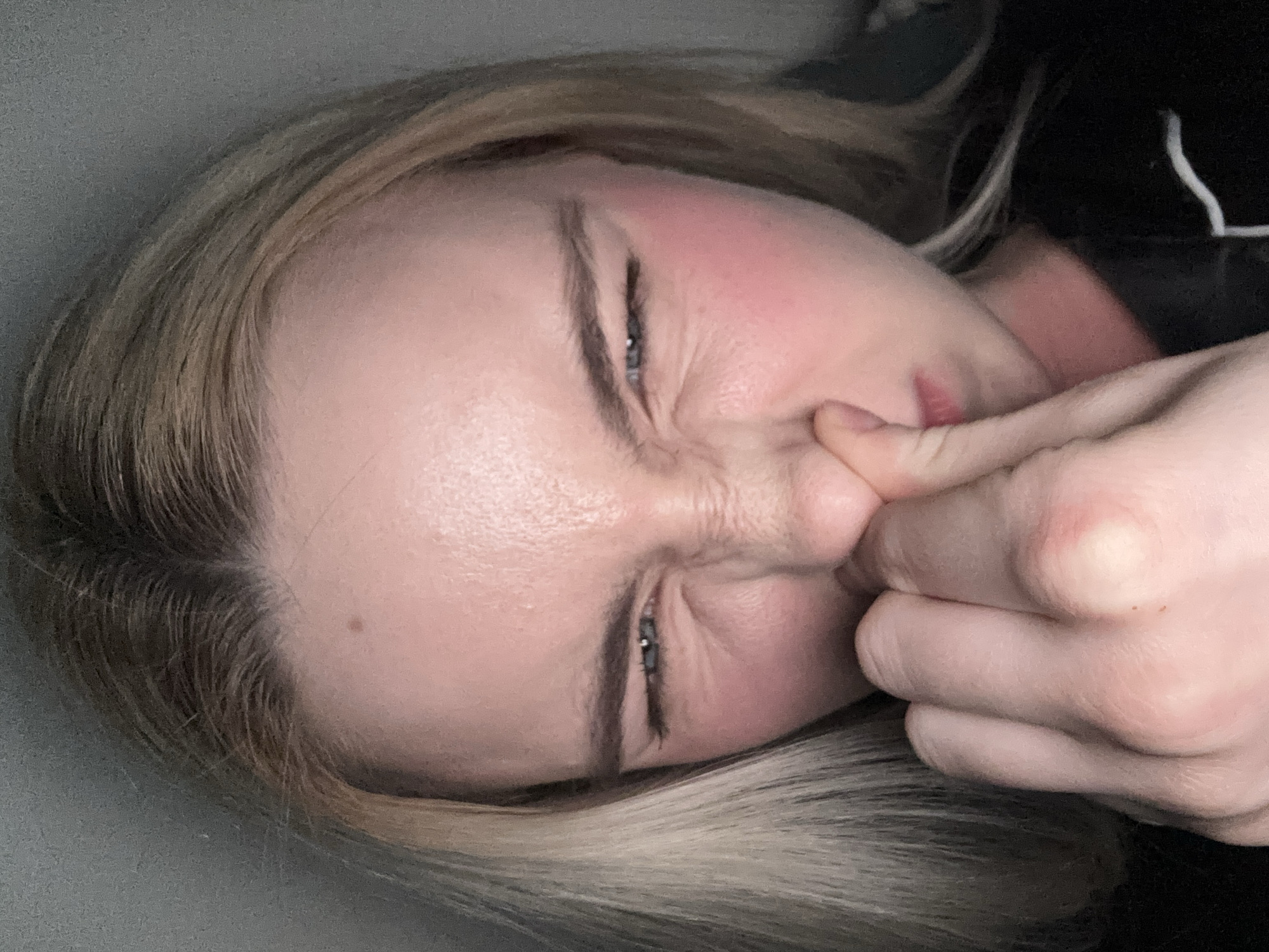 A close-up, angled portrait captures a woman pinching her nose with her fingers, her face deeply contorted in an expression of strong displeasure or disgust. Her eyes are squeezed tightly shut, and her brow is furrowed, causing prominent wrinkles around her nose and eye area. Her cheeks show a rosy flush. She has light-colored hair, possibly blonde or light brown, which appears slightly disheveled. A dark garment, featuring a distinctive white piping or stitching detail, is visible near her head and shoulder. The background is a plain, dark, undifferentiated surface. No text is visible in the image. This scene takes place in Vsevolozhsk, Russia.