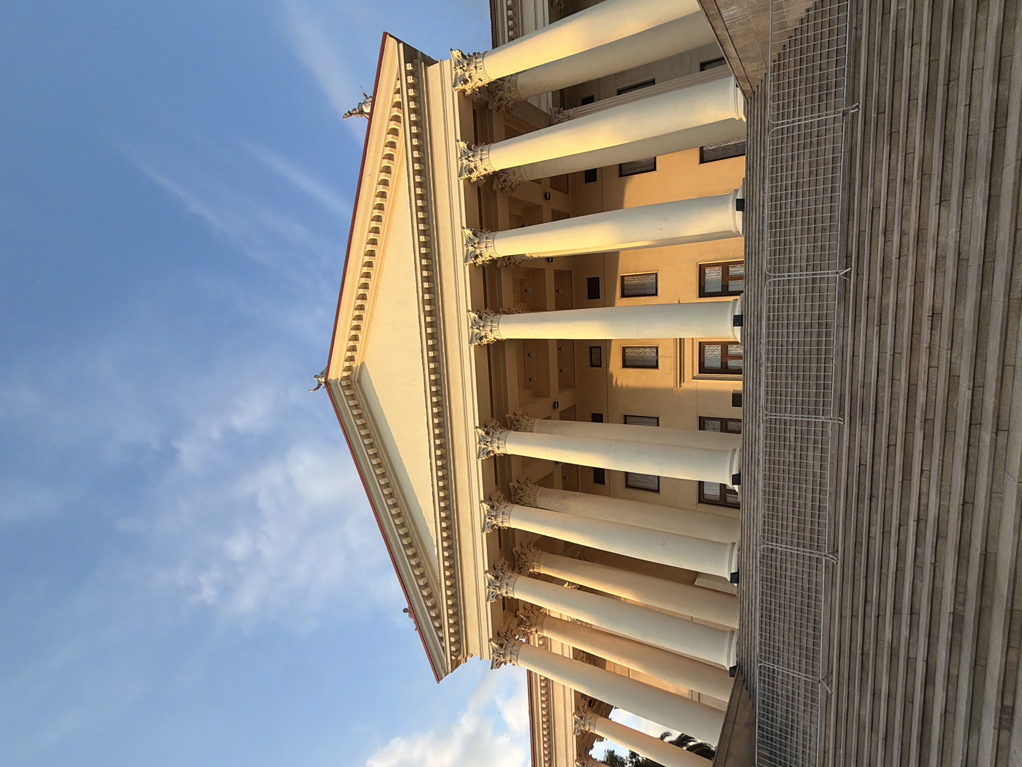 The image shows the facade of a grand, neoclassical building in Sochi, Russia, specifically the State Museum of the History of Religion. The building features a prominent portico with a series of large, fluted Ionic columns supporting a triangular pediment. The facade is painted a pale yellow, and the roofline is accented with a red trim. Two small, dark-colored statues of figures in classical attire are visible atop the pediment. The building is accessed by a wide, stone staircase, which is partially obscured by a temporary metal barrier at the top. The sky above is a clear blue with a few wispy white clouds, and the setting sun casts a warm, golden light on the building's columns and facade.