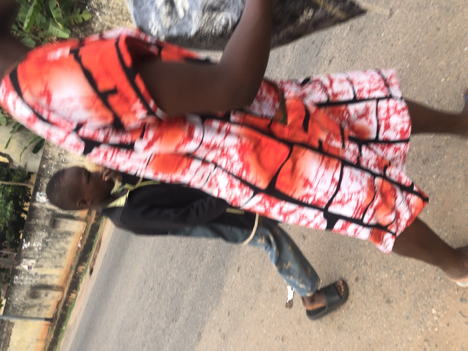 An image shows a child wearing a dark jacket, jeans, and sandals walking on pavement alongside a woman. The woman is wearing a short dress patterned with red, white, and black designs. The image is slightly blurry, and the background shows buildings and greenery. The shot appears to have been taken from an elevated viewpoint.