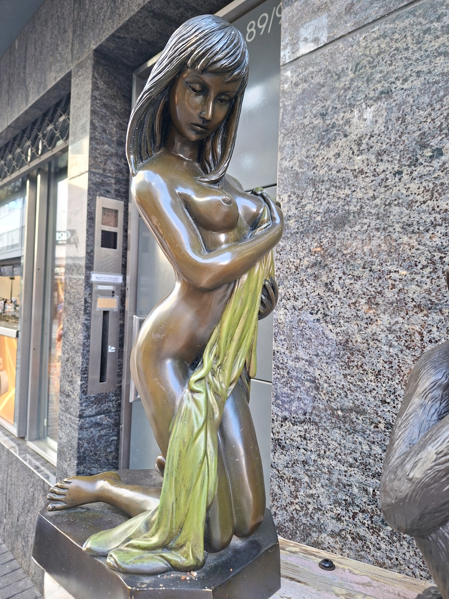 The image shows a bronze sculpture of a nude woman kneeling and holding a green cloth, in front of a building with a granite facade and glass windows. The sculpture is positioned on a dark base. A partially visible statue of a gorilla is located to the right of the woman sculpture. The building appears to be a commercial establishment, possibly a shop or an office building, given the visible signage "89/9" and a window display. The scene is outdoors, with paving stones visible in the foreground. The architectural style suggests an urban environment. Based on the presence of the "89/9" signage and the building's aesthetic, this sculpture is located on Hohenzollernring 89 in Cologne, Germany.