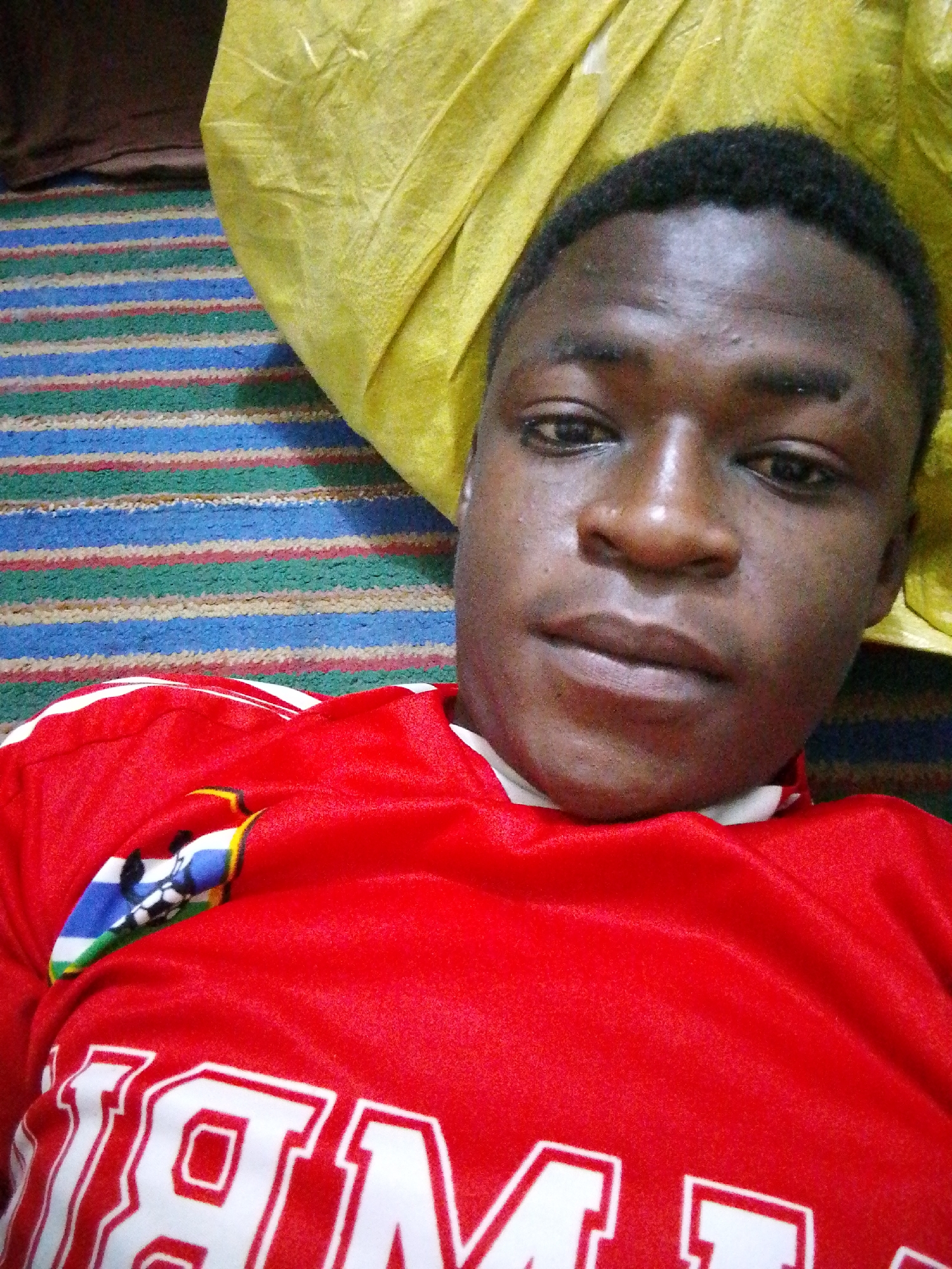 A young Black male is lying down, looking up at the camera. He is wearing a red shirt with white trim and some lettering, partially obscured by the frame. A crest featuring blue, white, green, and black colors is visible on the left side of his shirt. He has dark, short hair and a light brown complexion. He appears to be indoors, with a striped rug beneath him and a yellow fabric item positioned behind his head. The lighting is dim.