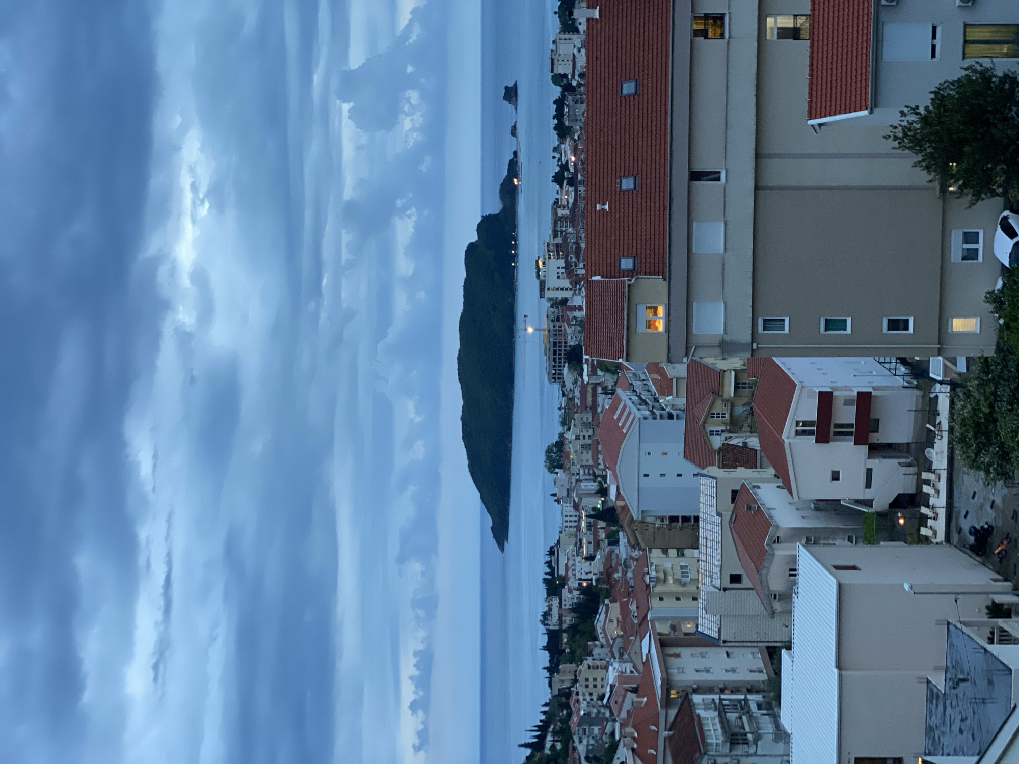 This elevated view captures a coastal town, likely Budva in Montenegro, at dusk or early night. The scene is dominated by a dramatic, overcast sky filled with heavy, blue-grey clouds that suggest impending rain or a recent storm. The horizon is low, with a calm, dark blue sea stretching across the midground.
On the left side of the image, a large, dark, tree-covered headland or island juts out into the sea, its silhouette prominent against the lighter water. Further out in the sea, a smaller, rocky islet is faintly visible. The coastline is dotted with lights from buildings and streetlights, creating a soft glow along the waterfront.
The town itself extends from the foreground to the midground, with numerous buildings featuring characteristic red-tiled roofs and light-colored facades. The architecture appears Mediterranean, with multi-story structures densely packed along the coast and climbing up the hillside. Many windows in the buildings display warm, artificial light, indicating occupancy and adding to the evening atmosphere. In the immediate foreground on the right, the top of a white car and some green foliage are visible, suggesting a street or parking area.
No people are visible, and there is no discernible activity beyond the general ambiance of an illuminated town at twilight. No text is visible in the image. The overall mood is serene yet slightly somber due to the dark, cloudy sky.