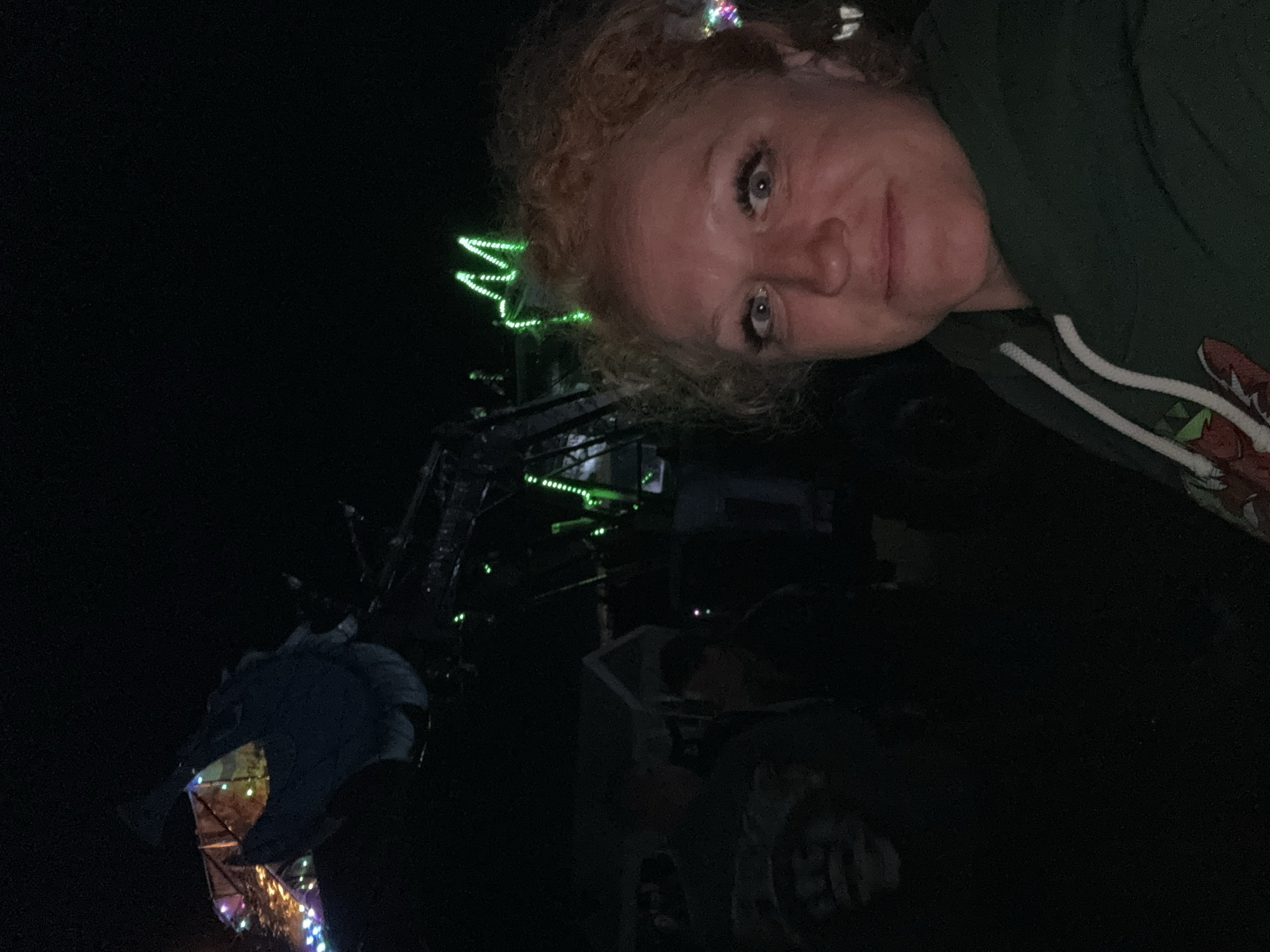The image depicts a nighttime scene in Большая Кивара, Russia, illuminated by artificial lights. In the foreground, a woman with light-colored, curly hair, light eyes, and prominent eyelashes faces the camera with a neutral to slight smile. She is wearing a dark green hooded sweatshirt with white drawstrings and a visible graphic on the lower left chest. Behind her, a large piece of dark-colored machinery, possibly agricultural equipment, is adorned with bright green LED strip lights, some forming a distinct pattern that resembles an 'M' or crown shape. To the left of the woman and the machinery, multicolored string lights (blue, green, yellow, purple) illuminate a fence or structure, and at least two additional indistinct human figures are partially visible in the darkness. The scene is otherwise dark, with minimal environmental details discernible beyond the illuminated elements.