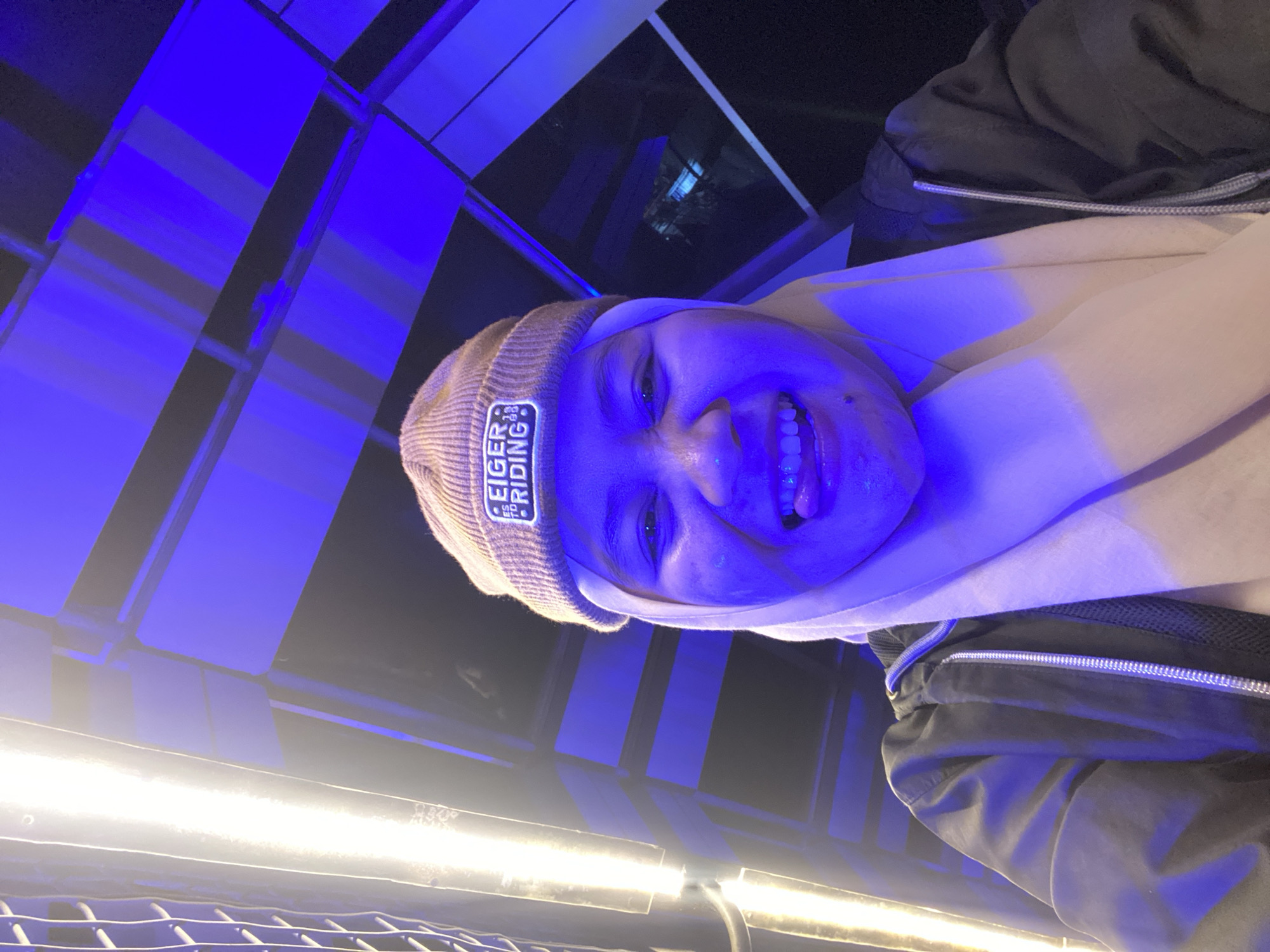 A person is captured in a selfie-style photograph, illuminated by strong blue and white lights. The subject is wearing a light-colored beanie with a patch that reads "EIGER RIDING." They are also wearing a light-colored hoodie or sweatshirt and a dark jacket. The person's face is close to the camera, with their mouth open and tongue visible, suggesting a playful or excited expression. The background is abstract, with panels of blue light and some darker structural elements, possibly indicating an indoor setting with dramatic lighting, like a stage, an entertainment venue, or a studio. There are also bright white lights in the foreground, adding to the artificial lighting environment. The overall impression is one of a candid and vibrant moment, possibly at night given the artificial lighting. The image is oriented sideways.