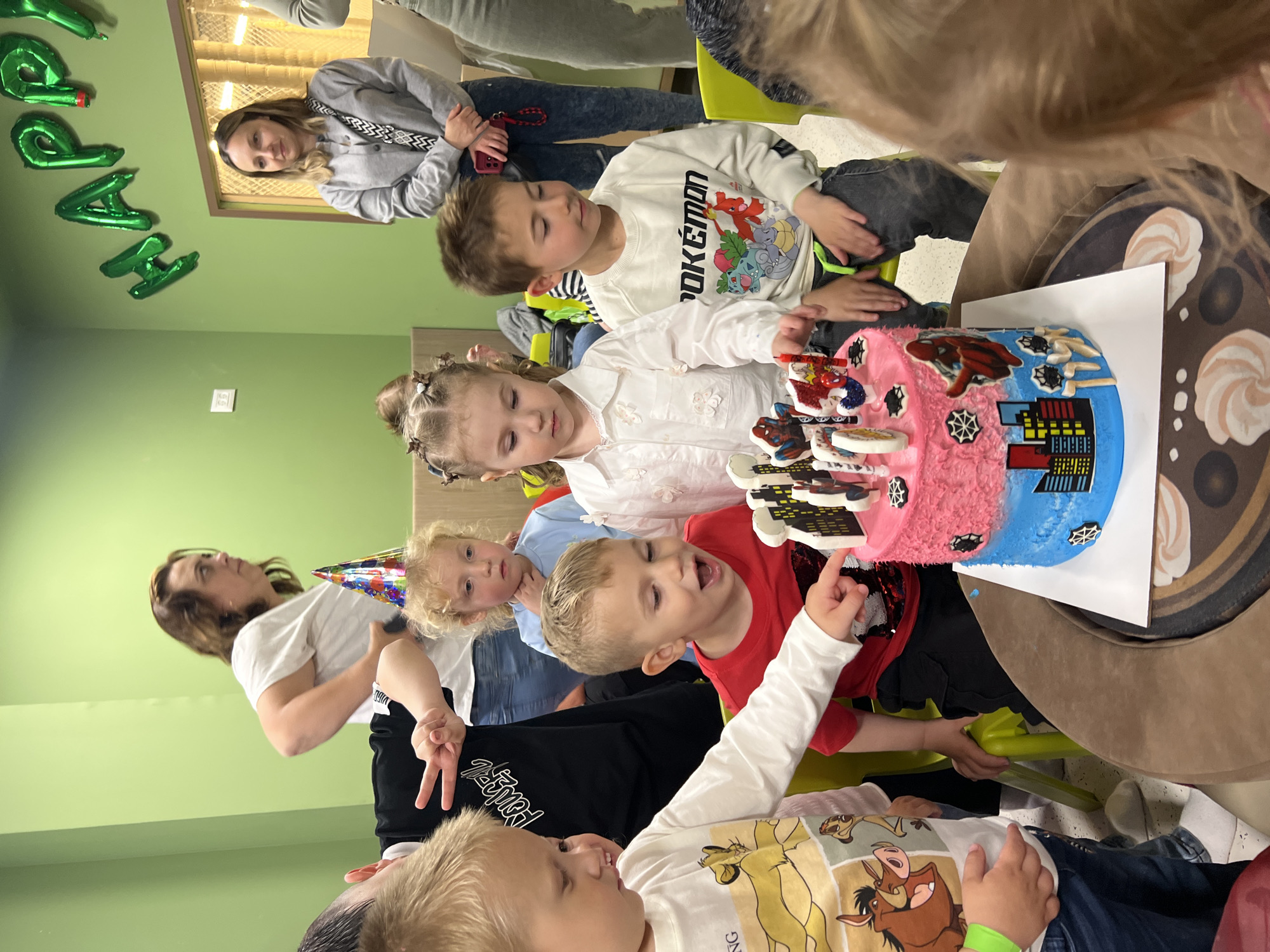 A group of children gather around a Spiderman-themed birthday cake. The cake is half pink and half blue, with a cityscape design on the blue half and Spiderman decorations on top. Several adults are also present, observing the scene. The environment is brightly lit with green walls, and balloon decorations with the word "HAPPY" are on one of the walls. The scene is a birthday celebration, taking place indoors, likely in a party room. It is not possible to confirm the location of Kiryat Bialik, Israel with the given information.