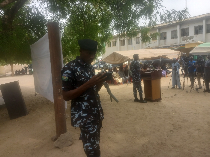 The image shows a scene in Maiduguri, Nigeria, at coordinates 11.869 latitude and 13.125 longitude. A man wearing a camouflage uniform and a green beret is standing on a dirt path, holding a black device in his hands. He is looking down at the device. Behind him, a large white banner hangs from a wooden pole. To the right of the man, a group of people are seated on chairs in front of a wooden lectern under a canopy tent. A person, seemingly a journalist, stands behind the lectern with a microphone in their hand, and several other people are standing behind the lectern, holding cameras and microphones. The background includes a building with windows, tents, and cars. There is a tree on the left side of the image. The scene appears to be an event, potentially a press conference or a public gathering.