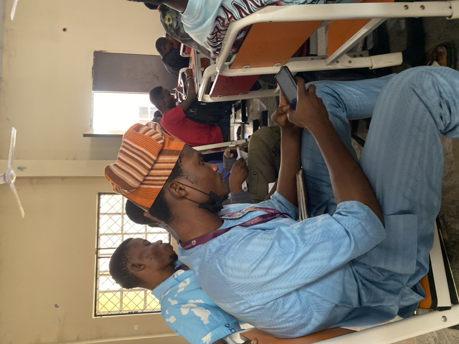 The scene is set inside a classroom or lecture hall in Minna, Nigeria, occupied by multiple individuals, primarily young adult males, seated at desks.
In the foreground, a dark-skinned male is seated in profile, facing right, on an orange chair with a white metal frame. He is dressed in a light blue traditional Nigerian long-sleeved top and matching trousers with a subtle patterned texture. A multi-colored (orange, brown, and beige striped) cap adorns his head. A black face mask is positioned below his chin, and a maroon lanyard with partially visible white text hangs around his neck. He is actively holding a black smartphone with both hands, his gaze fixed on its illuminated screen.
Behind the foreground subject, another dark-skinned male is visible, wearing a light blue short-sleeved shirt with white cloud-like patterns, also looking downwards. Further into the midground, other individuals are seated at similar desk-chair units with white frames and orange or red seating. One individual in a red t-shirt is visible with a black backpack, and others are seen looking down, possibly engaged in writing or similar activities.
The room's walls are painted a faded light yellow or beige, showing signs of wear, including stains and minor surface damage. Two windows with horizontal metal security bars are present on the left side, admitting bright natural light. A white four-bladed ceiling fan is mounted overhead. A partially open doorway on the right side of the frame reveals a brightly lit area beyond. The floor is a dark, unadorned surface.