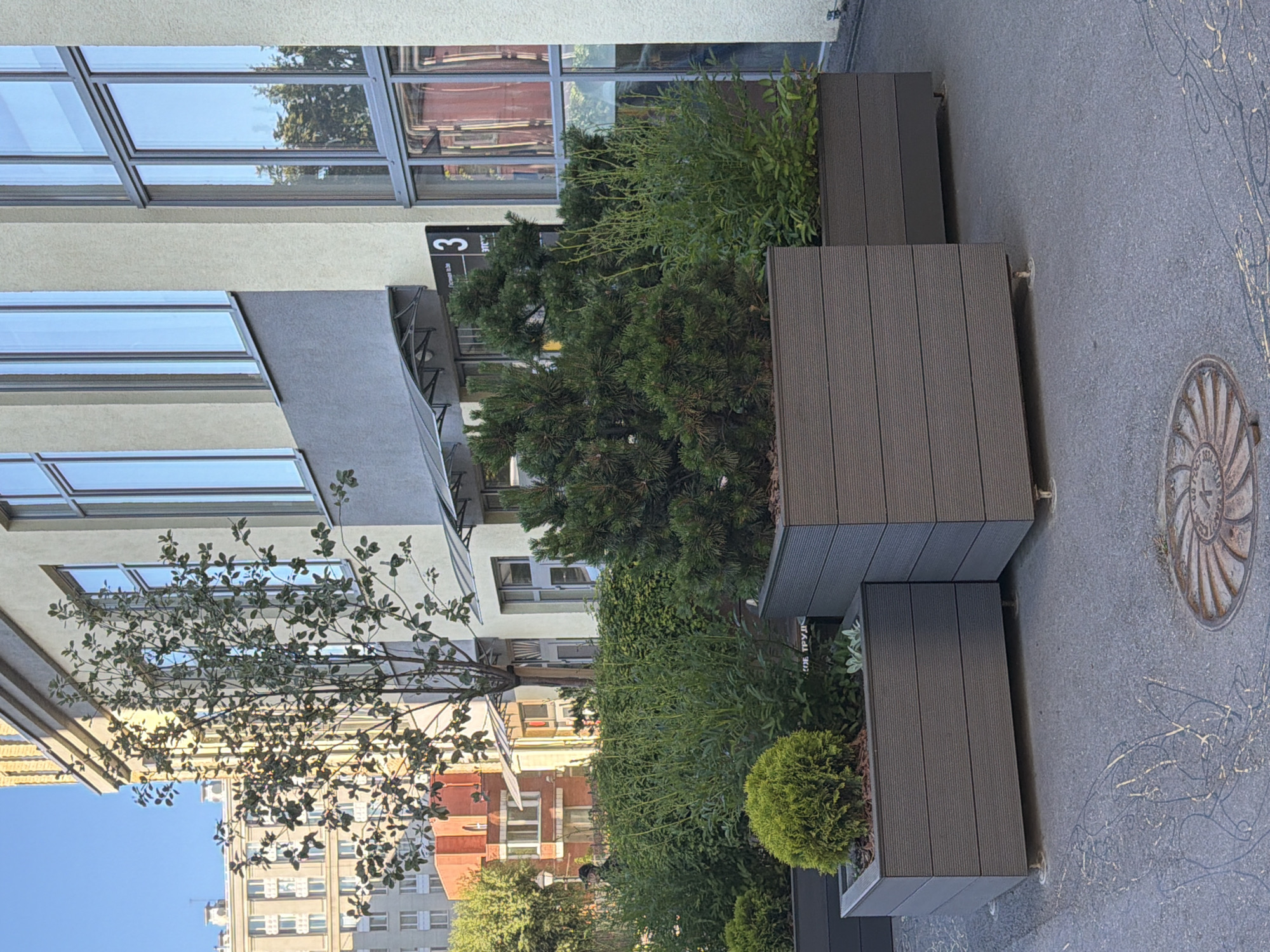 In Saint Petersburg, Russia, on a paved city sidewalk, a series of large, modern, dark gray planters are arranged. The planters are rectangular, constructed of horizontally slatted material, and elevated slightly off the ground by small metal feet. The planters contain various greenery. One planter features a dense, spherical evergreen shrub and other smaller plants. Another planter holds a bushy pine tree with dark green needles. A third, longer planter contains a tall, bushy green plant with thin, light green stems and small leaves. Behind the planters, a light-colored building with large, floor-to-ceiling windows dominates the scene. A smaller, gray awning is situated above a portion of the building's entrance. A black sign with a white number "3" and some text is affixed to the building's facade. To the left of the building, a tall tree with sparse leaves casts a shadow. Further in the background, a multi-story building with a beige facade and many windows is visible. A person, partially obscured by foliage and wearing dark clothing, can be seen walking to the left of the planters. The ground in front of the planters is asphalt, with decorative linear markings and a circular metal manhole cover. The lighting suggests it is daytime, with sunlight illuminating the scene.