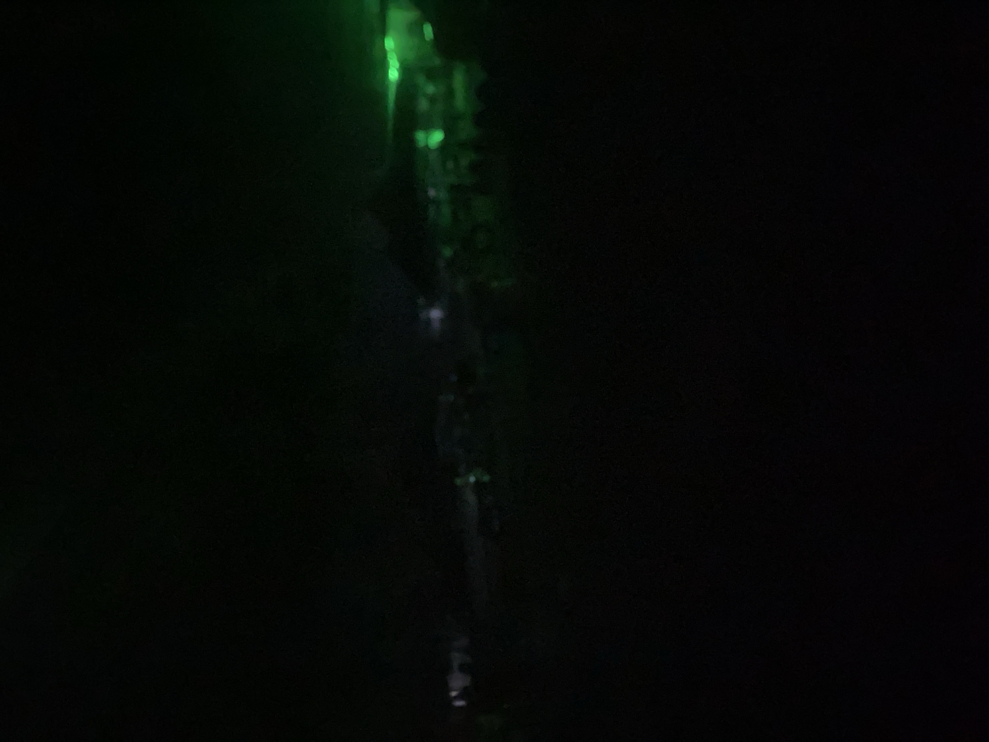 The image depicts a dimly lit scene, possibly an outdoor event or gathering, in Howayi, Nigeria. The majority of the image is in shadow, with the exception of some areas illuminated by green lighting. These lights highlight indistinct shapes that could be structures or a crowd of people. The green light emanates from the right side of the image, casting a glow over the middle ground. The overall impression is one of low visibility and an event taking place at night.