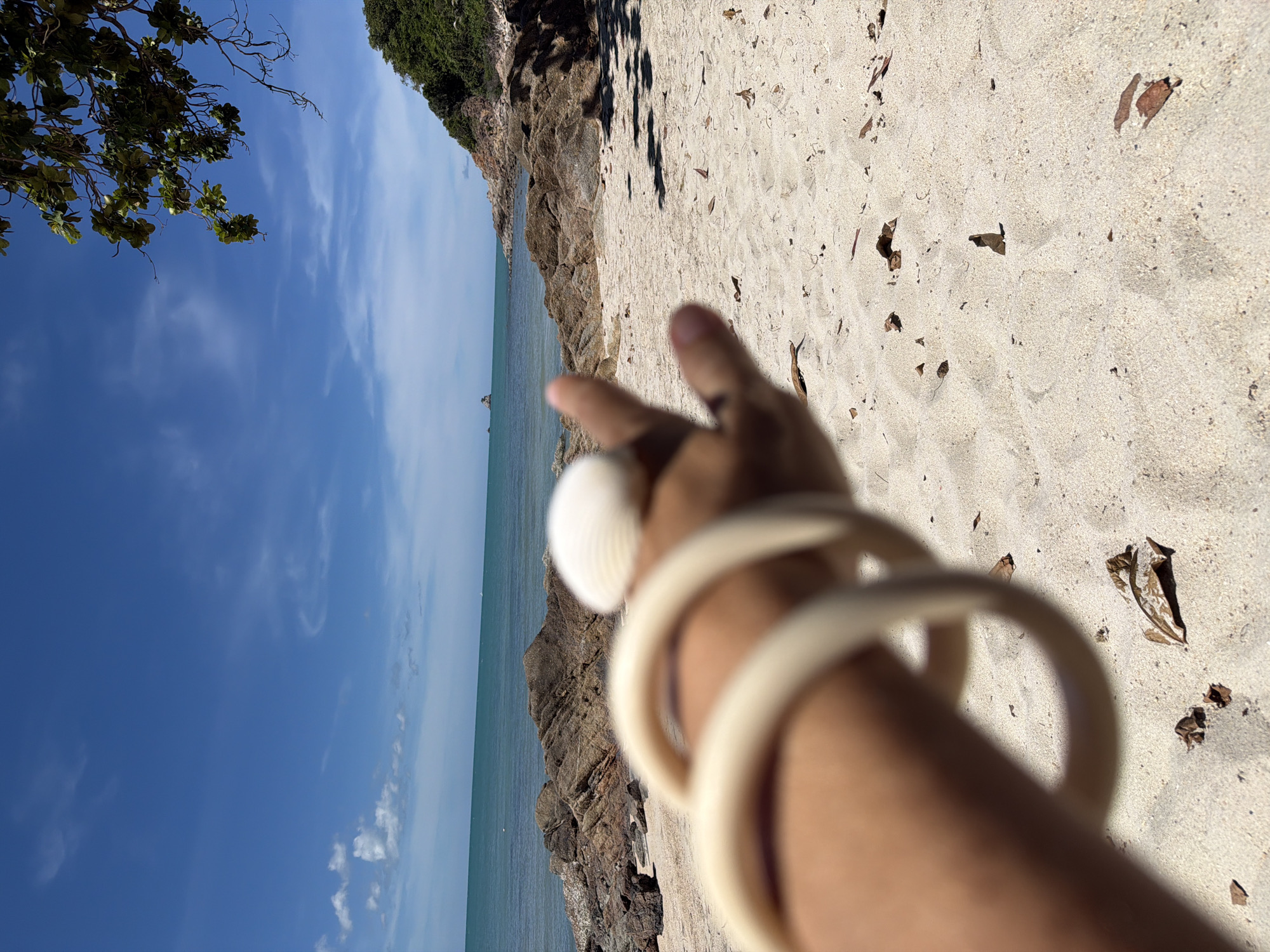 This is a close-up, low-angle, vertical shot of a person's arm and hand at a beach in Baan Plai Laem, Thailand. The person is holding up a seashell and making a peace sign with their fingers. The arm is adorned with a light-colored bangle or bracelet.
The background features a sandy beach with scattered dry leaves and debris. To the left, there are rocky outcrops leading to the ocean. The sea is a calm, turquoise color, and the sky is a clear blue with wispy white clouds. The sun appears to be high in the sky, suggesting it is midday. The scene evokes a sense of relaxation and enjoyment of a tropical environment. No visible text is present in the image.