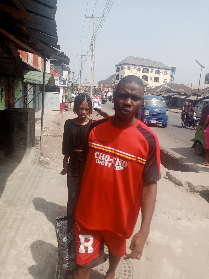 The image depicts a street scene in Aba, Nigeria, under a bright, hazy sky. In the immediate foreground, a dark-skinned male faces the camera. He wears a red and black short-sleeved athletic shirt with "SPORT" on the left shoulder and "CHO-CHO UNITY CUP" emblazoned across the chest. He also wears red shorts with a white capital "R" on the left leg. A dark, possibly woven bag with faint white lettering ("SHO") hangs from his left shoulder. Behind him and slightly to his left, a dark-skinned female walks. She has braided hair and wears a black long-sleeved top and dark patterned trousers, holding a dark smartphone in her right hand.
To the left of the female, a large, dark rectangular concrete block sits on the concrete sidewalk. Buildings with corrugated metal roofs and awnings line the left side of the street. Numerous overhead power lines are strung between wooden and concrete utility poles that extend down the street. The road surface is visible, and a blue minivan or small truck is positioned in the midground on the right side of the street. Further down the street, several individuals are visible, including at least one person riding a motorcycle, with a second individual as a passenger. A multi-story building with numerous windows stands in the background on the right. A circular metal manhole cover is visible on the ground near the foreground male's feet. The overall scene suggests a busy commercial or residential street.