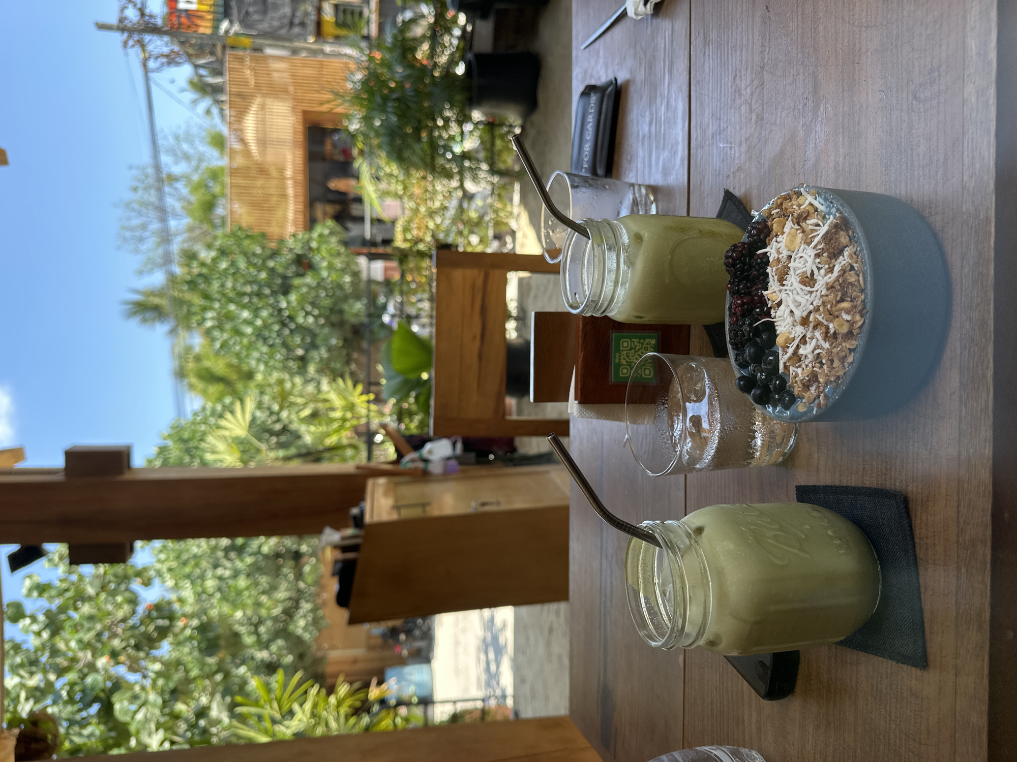 The image shows a wooden table in an outdoor setting, likely a cafe or restaurant. There are two mason jar glasses filled with a pale green liquid, possibly smoothies or juice, each with a stainless steel straw. Between them are two clear glasses, one containing water. To the right is a blue bowl filled with what appears to be a mix of granola, shredded coconut, and various berries. Each glass is placed on a square black coaster. The background includes greenery, suggesting a tropical environment, and parts of wooden structures. People can be seen in the distant background, but they are not clearly identifiable. The image is most likely taken in Puerto Escondido, Mexico.