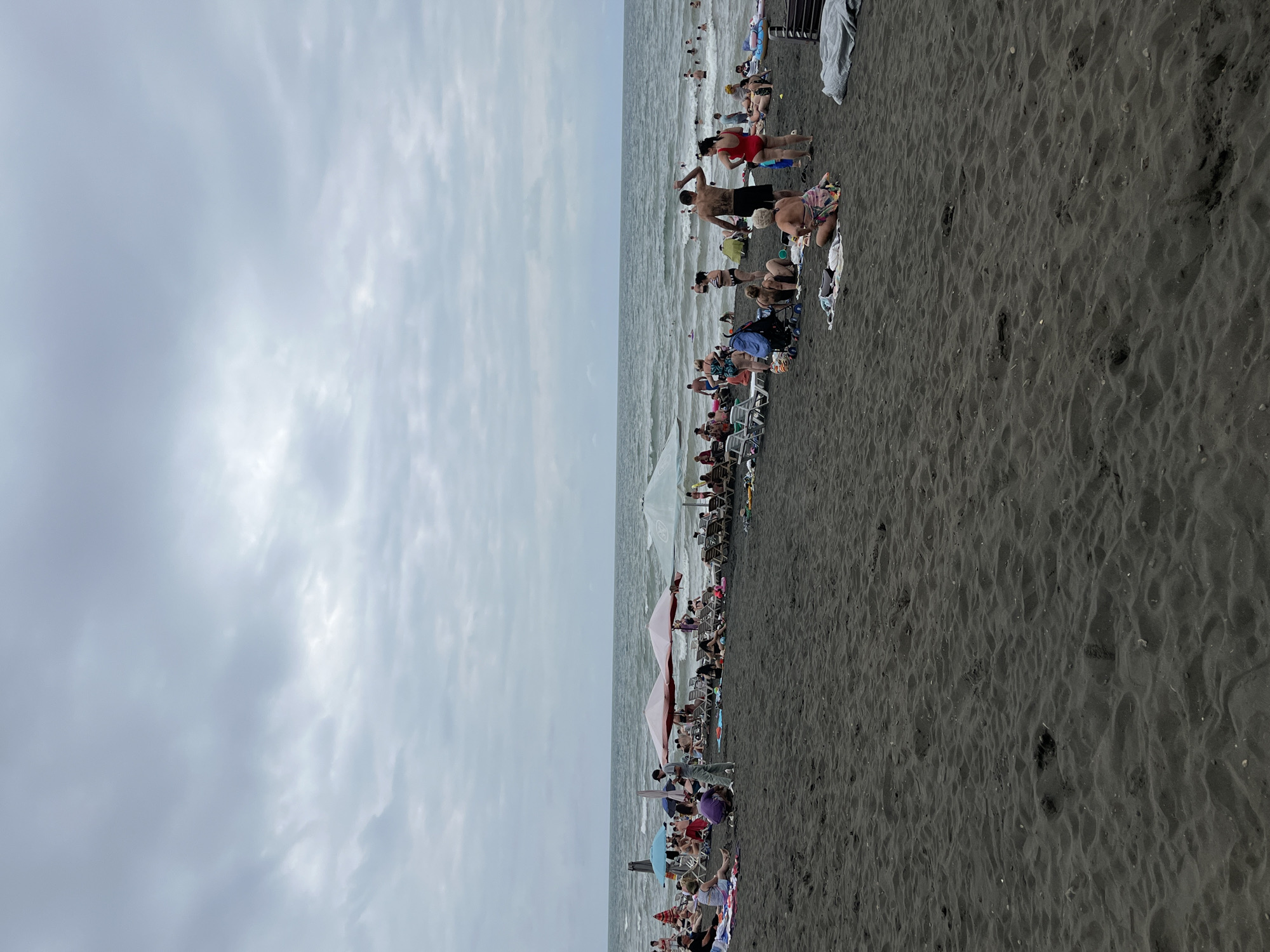 The image depicts a crowded black sand beach in Ureki, Georgia. The sky is overcast with gray clouds. People are spread across the sand, some sitting on towels and loungers, others standing or walking. Several beach umbrellas are set up. In the background, the sea is visible with moderate waves, and numerous individuals are in the water, swimming or wading. A few beach chairs and a stroller are also present on the sand. The dark sand covers the foreground and stretches to the water's edge.